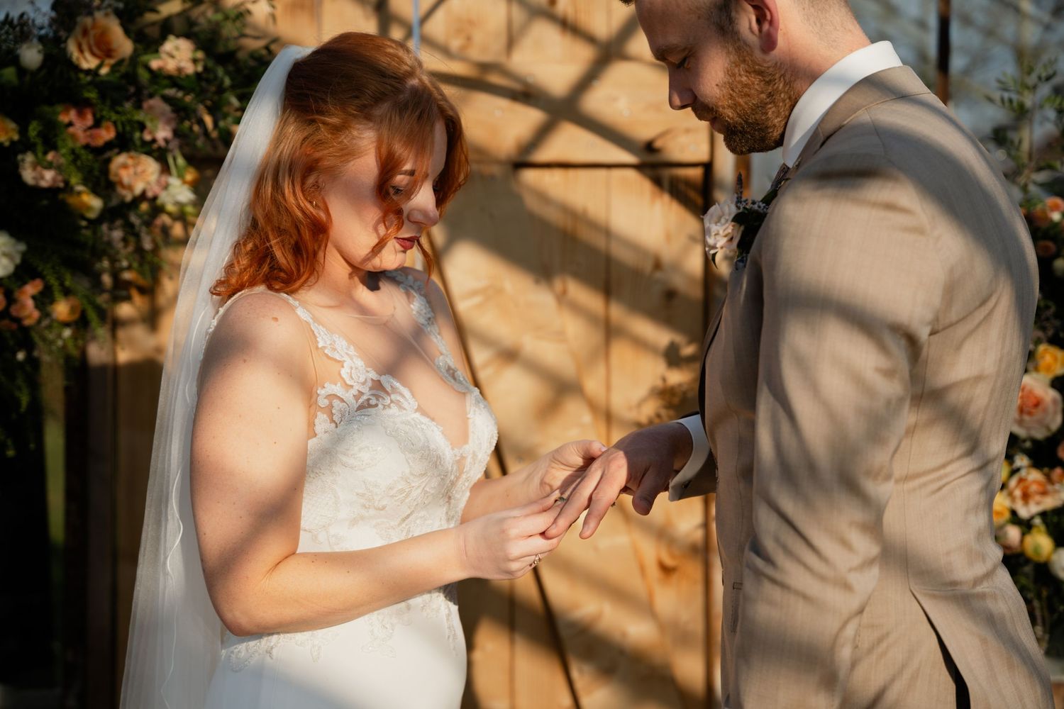 Bride placing wedding ring on groom's finger during outdoor ceremony with floral backdrop in warm sunlight.