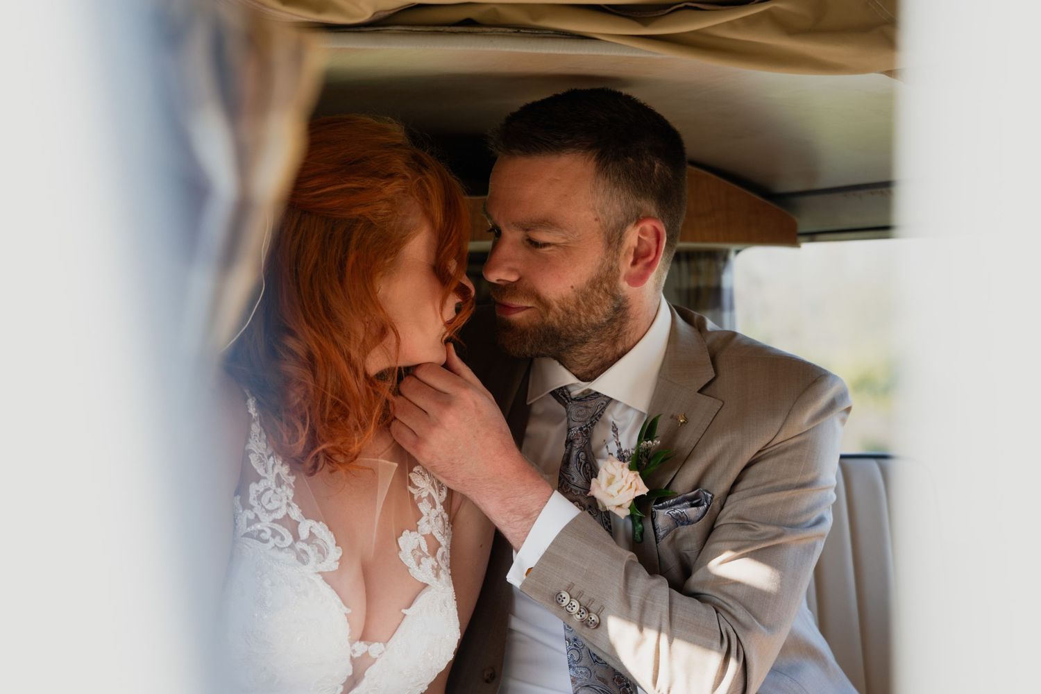 A groom in a gray suit tenderly holds his red-haired bride's face as they share an intimate moment inside a vintage car.