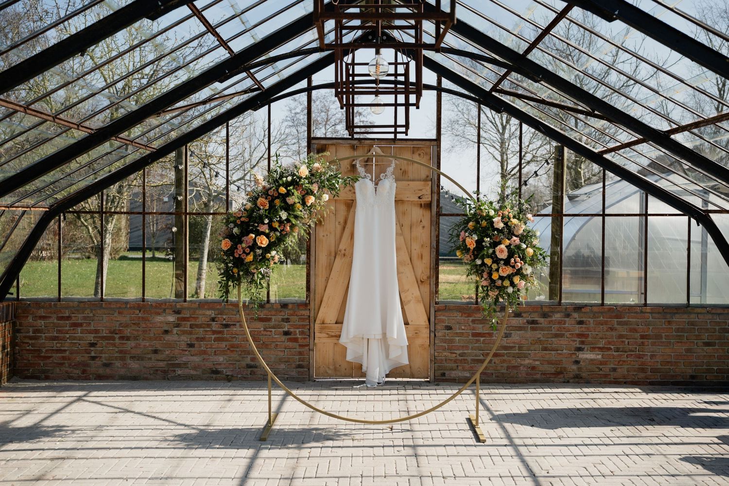 Wedding dress displayed on a circular floral arch inside a rustic greenhouse with brick walls and glass ceiling.