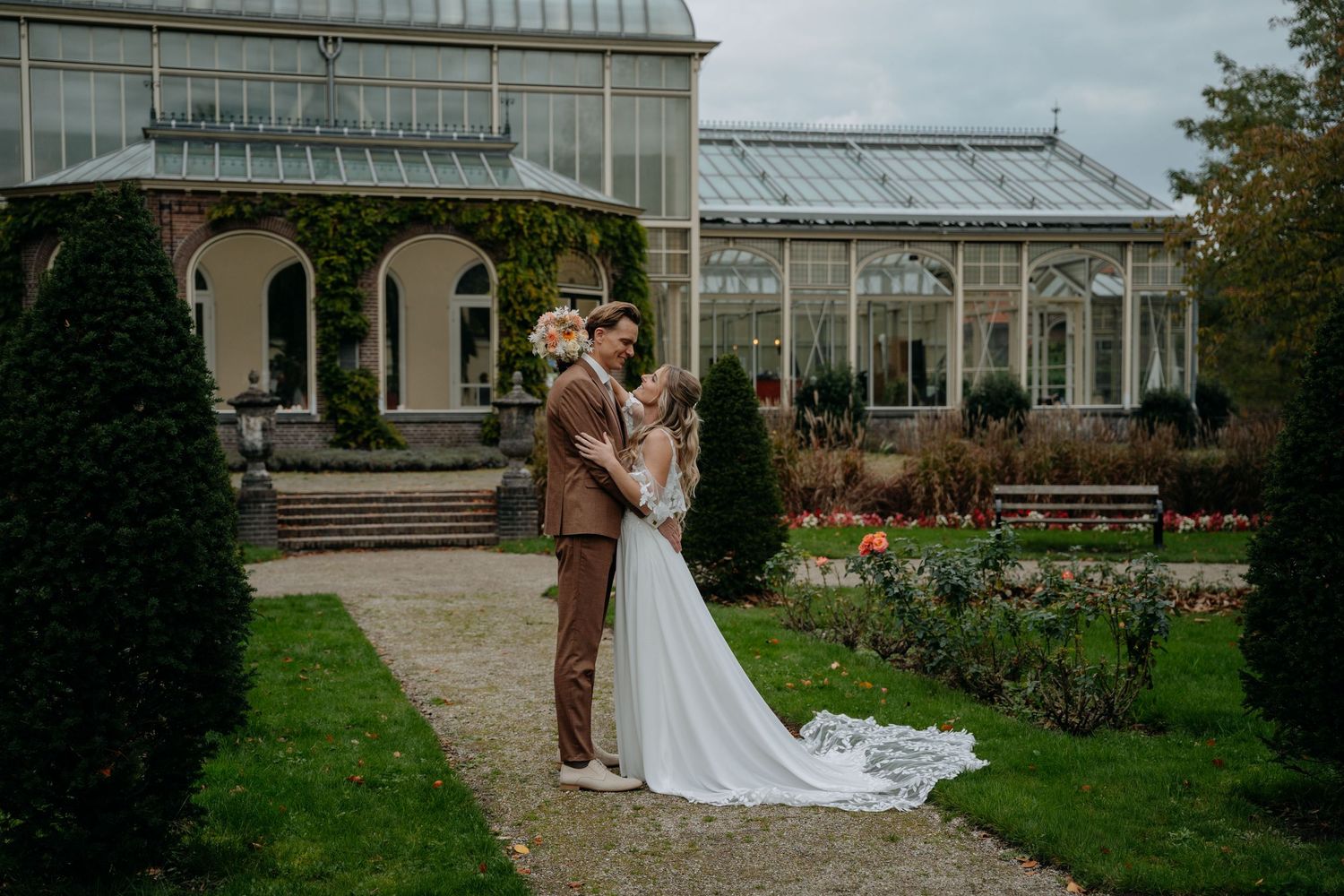 Bride and groom embrace in a lush garden with a stunning glass greenhouse backdrop on their wedding day.