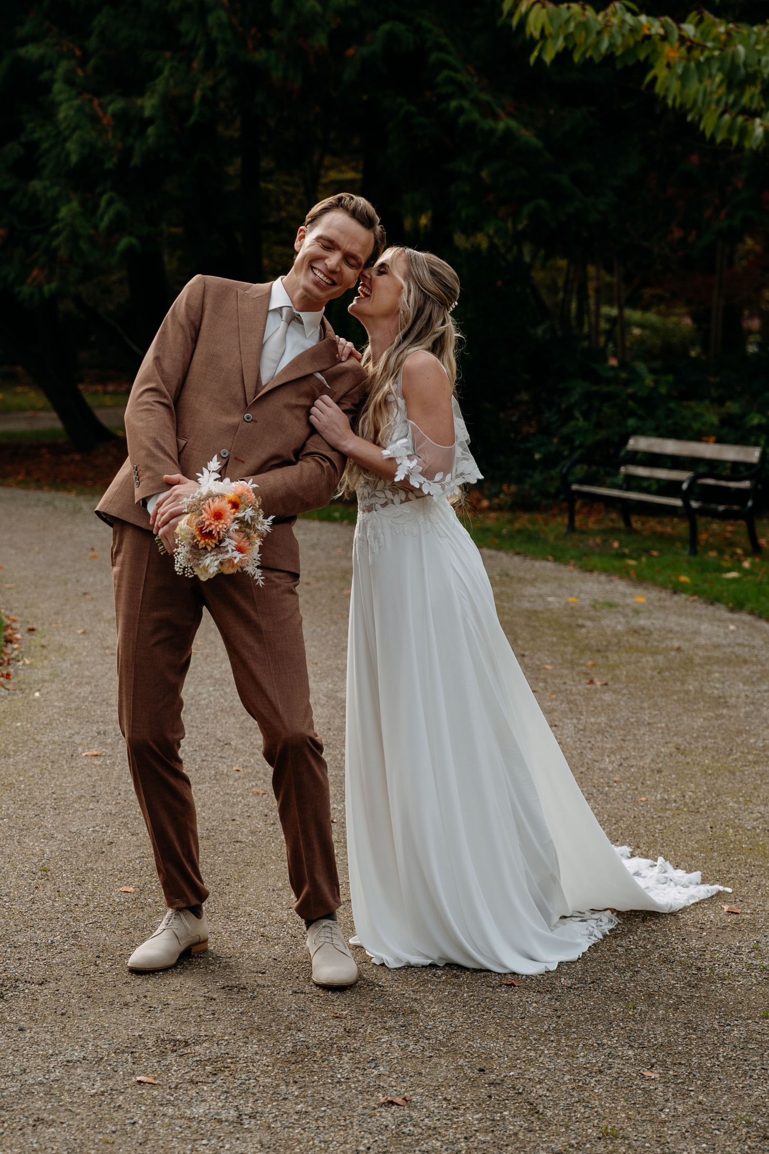 A couple in wedding attire share a romantic moment on a gravel path with autumn trees in the background.