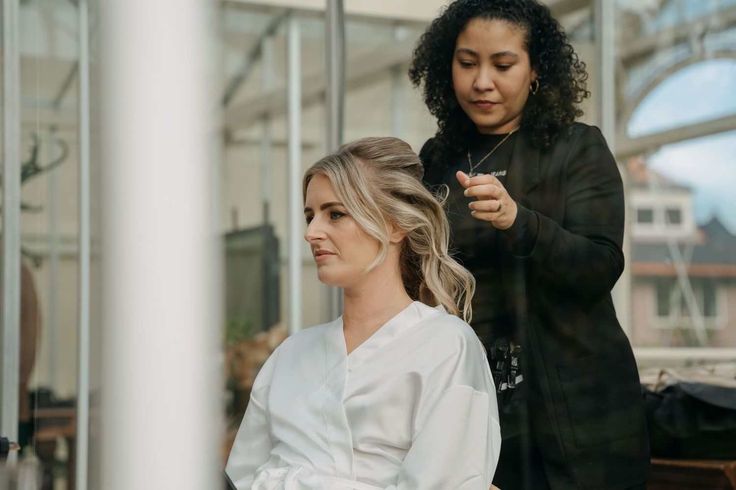 A hairstylist in black works on a client wearing a white robe in a salon setting.