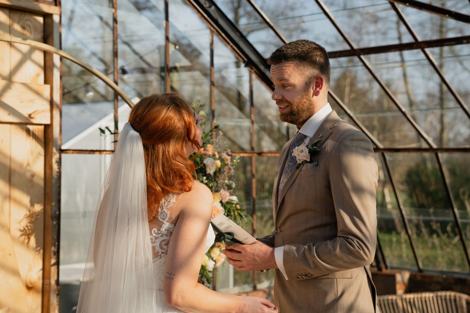 Bride and groom exchange vows in a sunlit greenhouse wedding ceremony, surrounded by floral greenery.