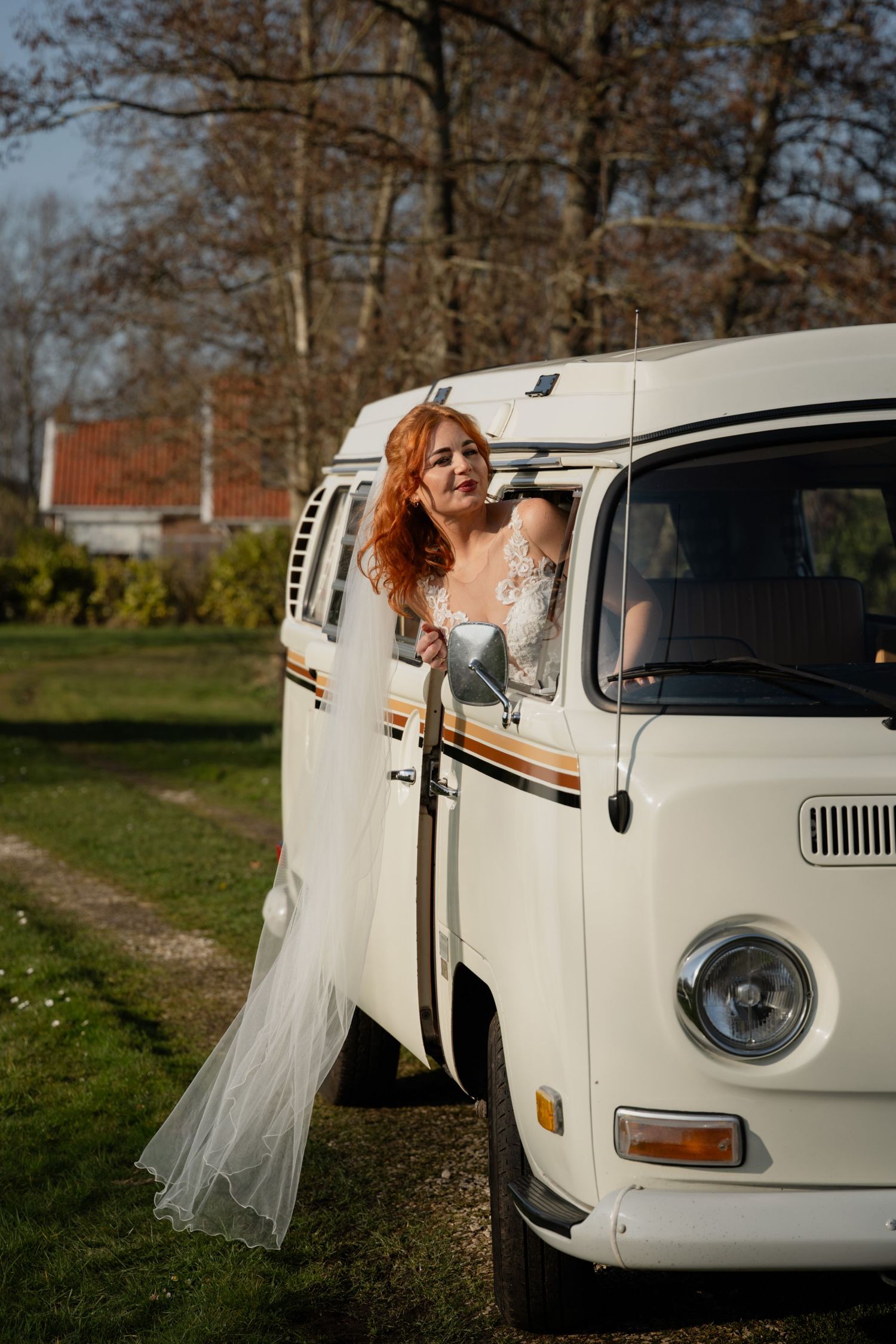Red-haired bride in white lace gown posing beside a vintage VW camper van outdoors on a sunny day.