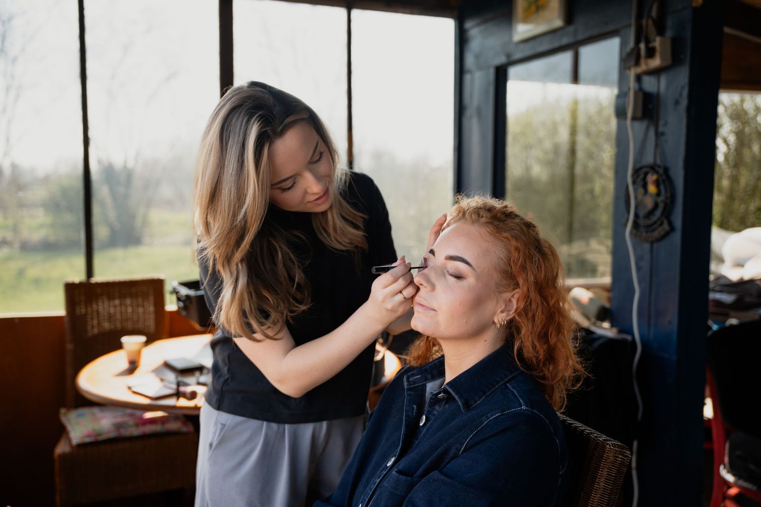 Makeup artist applying eyeshadow to a redhead woman in a bright studio with large windows.