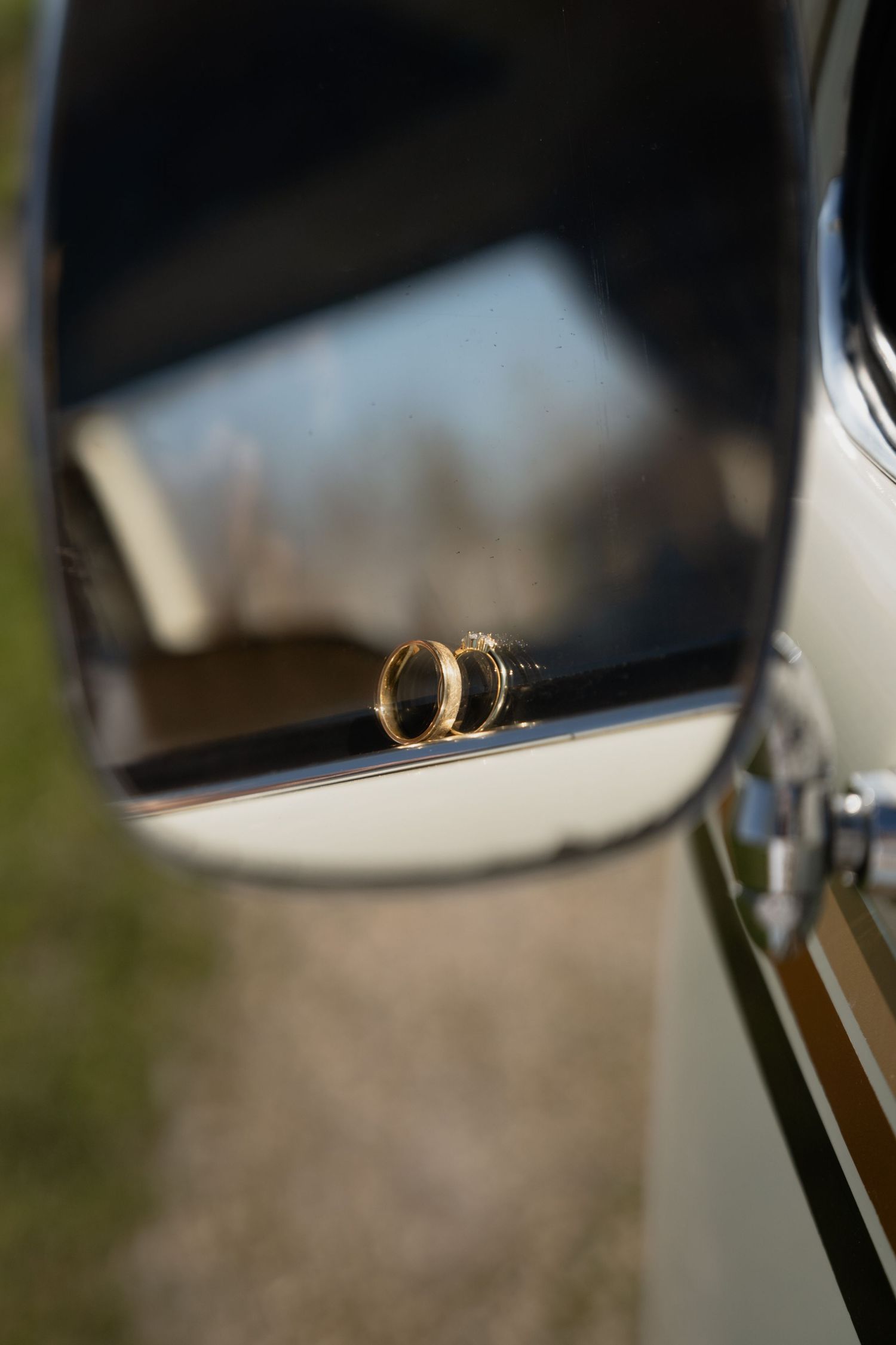 Two gold wedding rings resting on a vintage car side mirror, reflecting a blurred outdoor background.