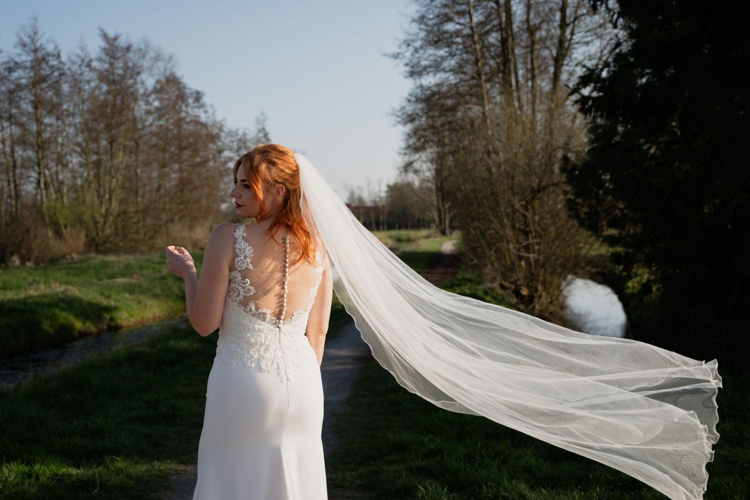 Bride in elegant backless wedding dress with flowing veil blowing in wind, standing in green outdoor setting.