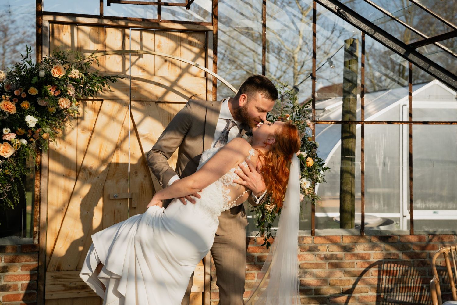 Groom dips bride in romantic kiss outside greenhouse wedding venue with colorful floral arch and warm sunlight.