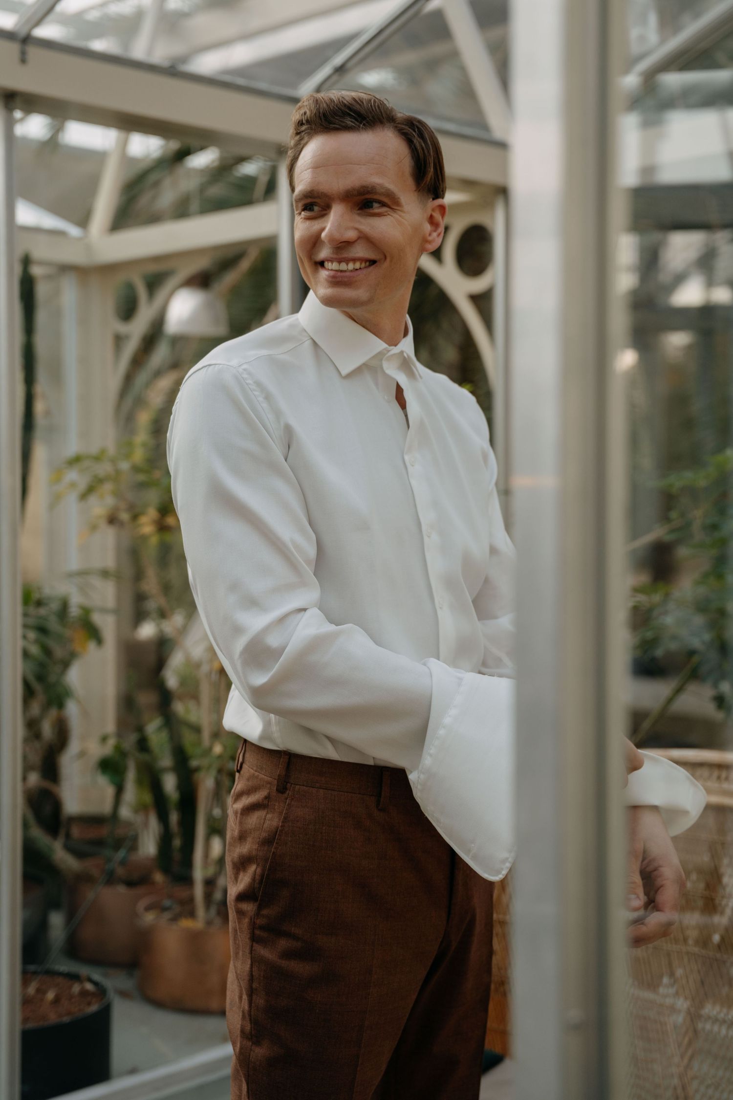 People in white shirts and brown pants stand in a bright conservatory or greenhouse with glass walls and plants.