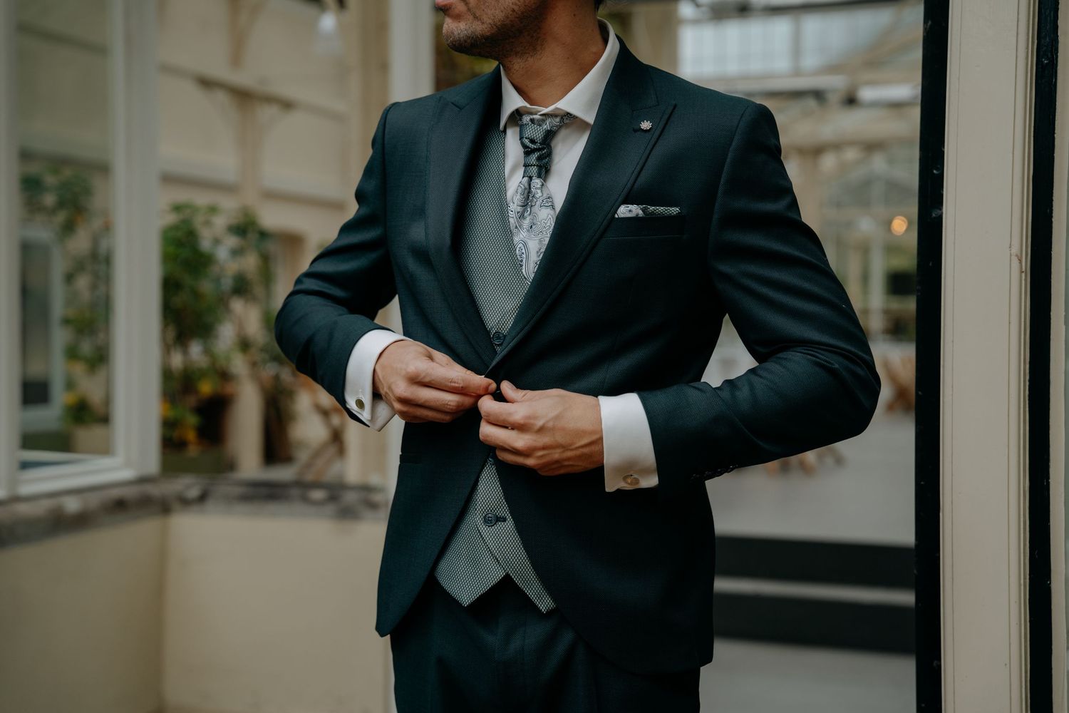Elegant groom adjusting suit jacket in sunlit venue hallway.