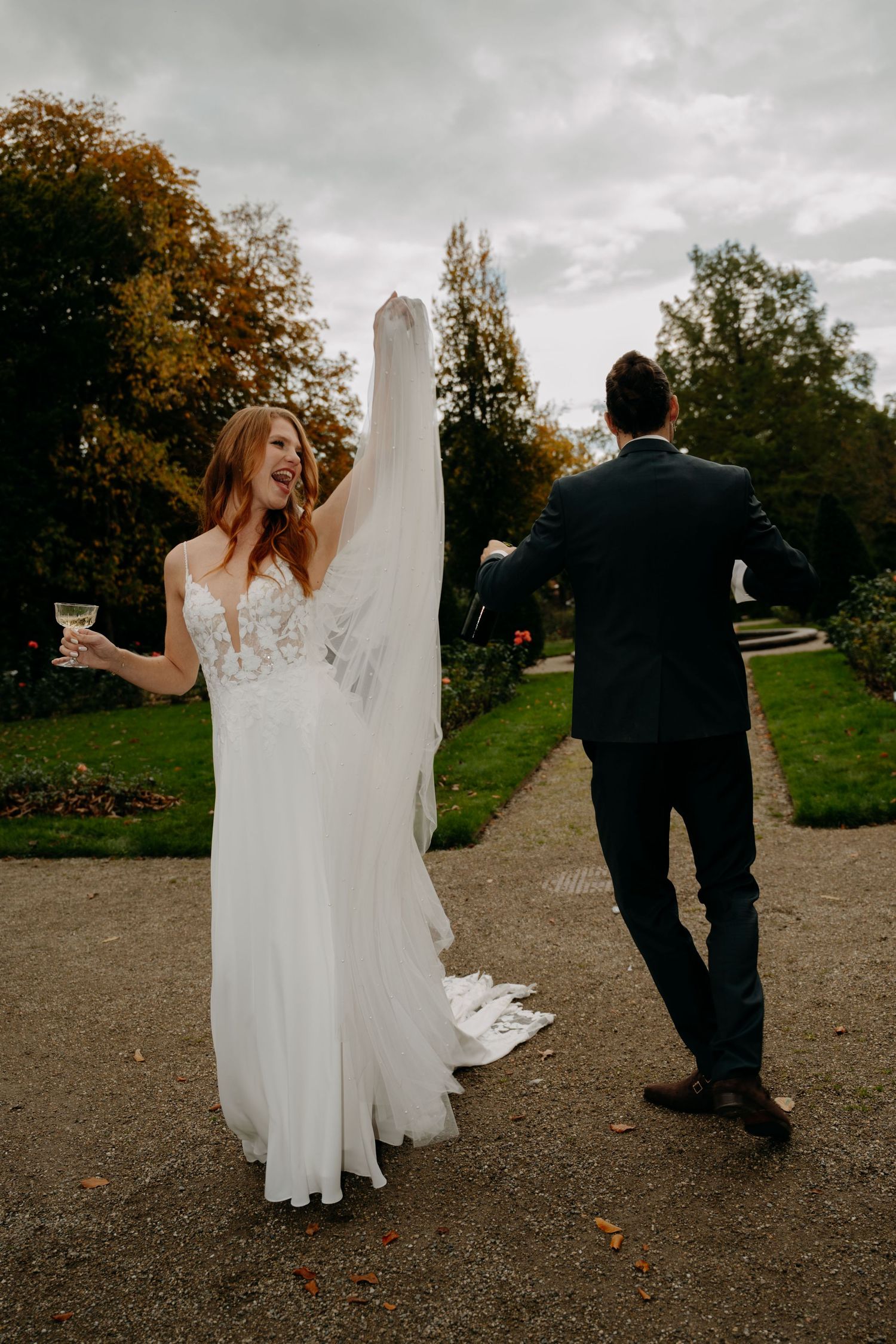 Bride in white lace gown holds veil and champagne while groom in navy suit walks ahead in garden.