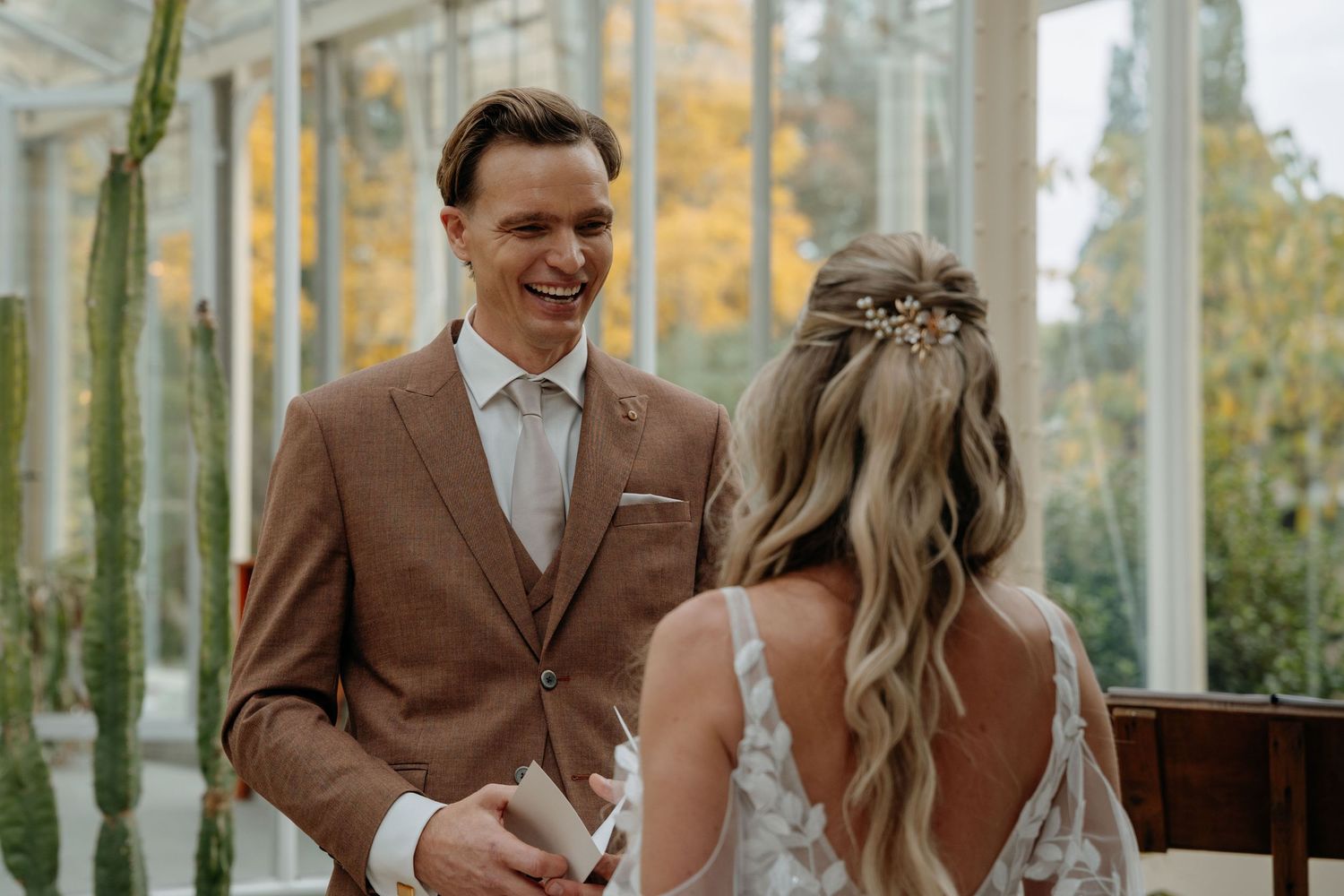 Wedding party in brown suits and light colored dresses gather outdoors during autumn with trees in background.