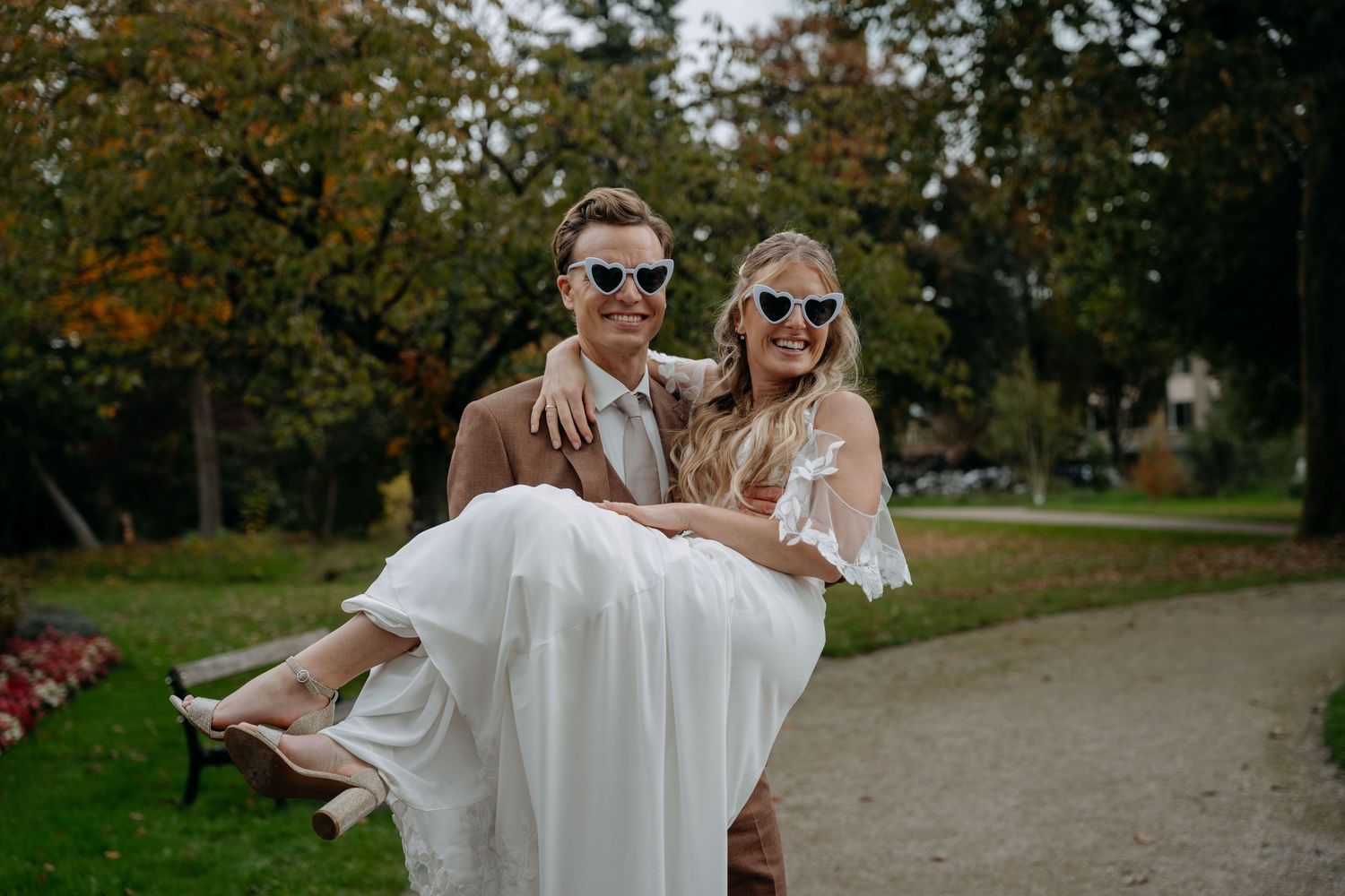 Smiling groom carries bride in white dress, both wearing sunglasses, in a lush green park on their wedding day.