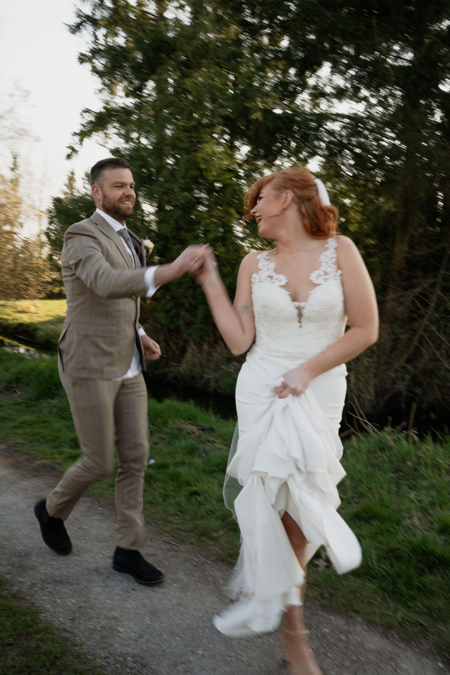 A joyful bride in a white lace gown and groom in a tan suit hold hands while running along a garden path.
