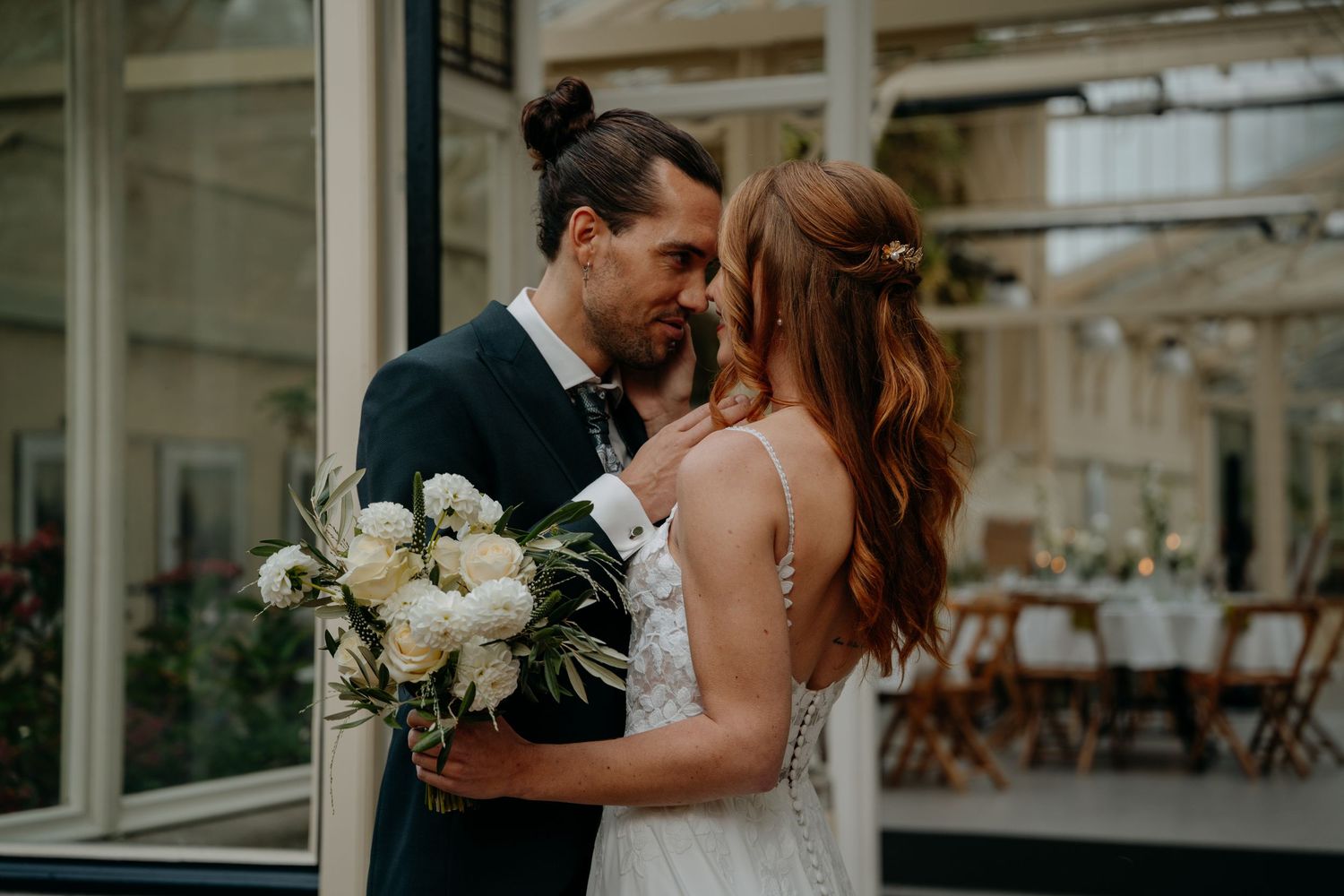 Elegant wedding couple embracing in front of a greenhouse-style venue.