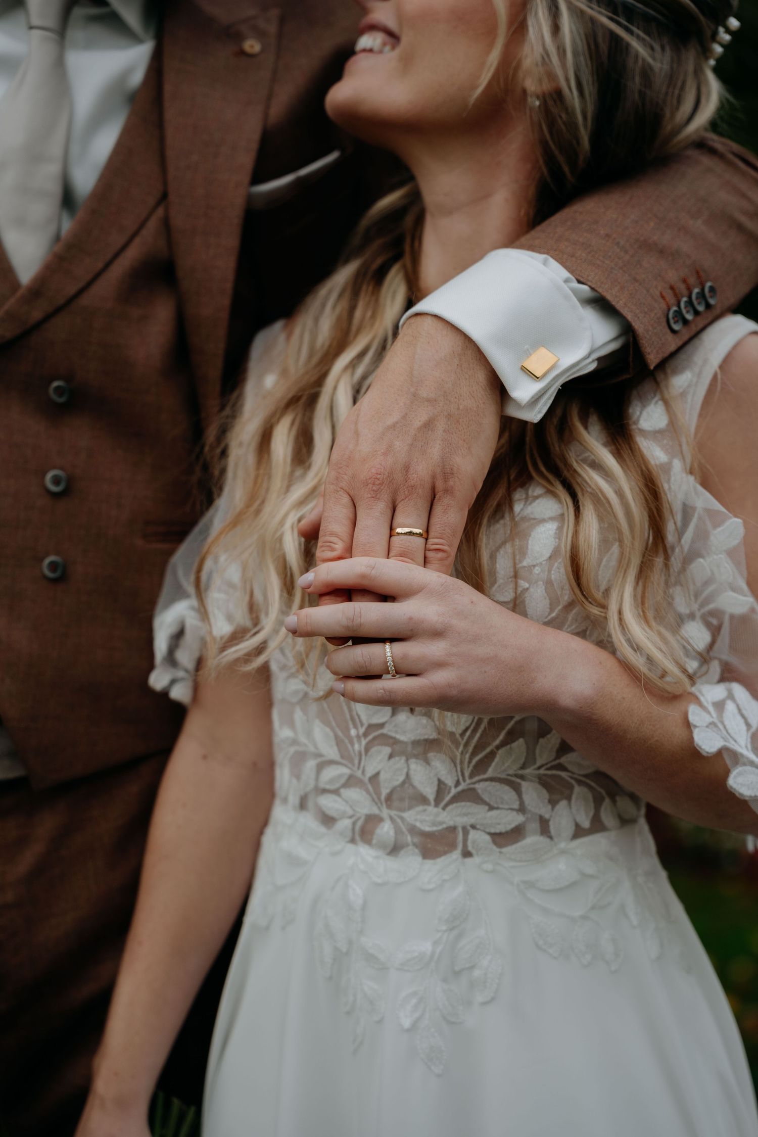 Close-up detail of a white lace wedding dress and brown suit with hands showing wedding rings.