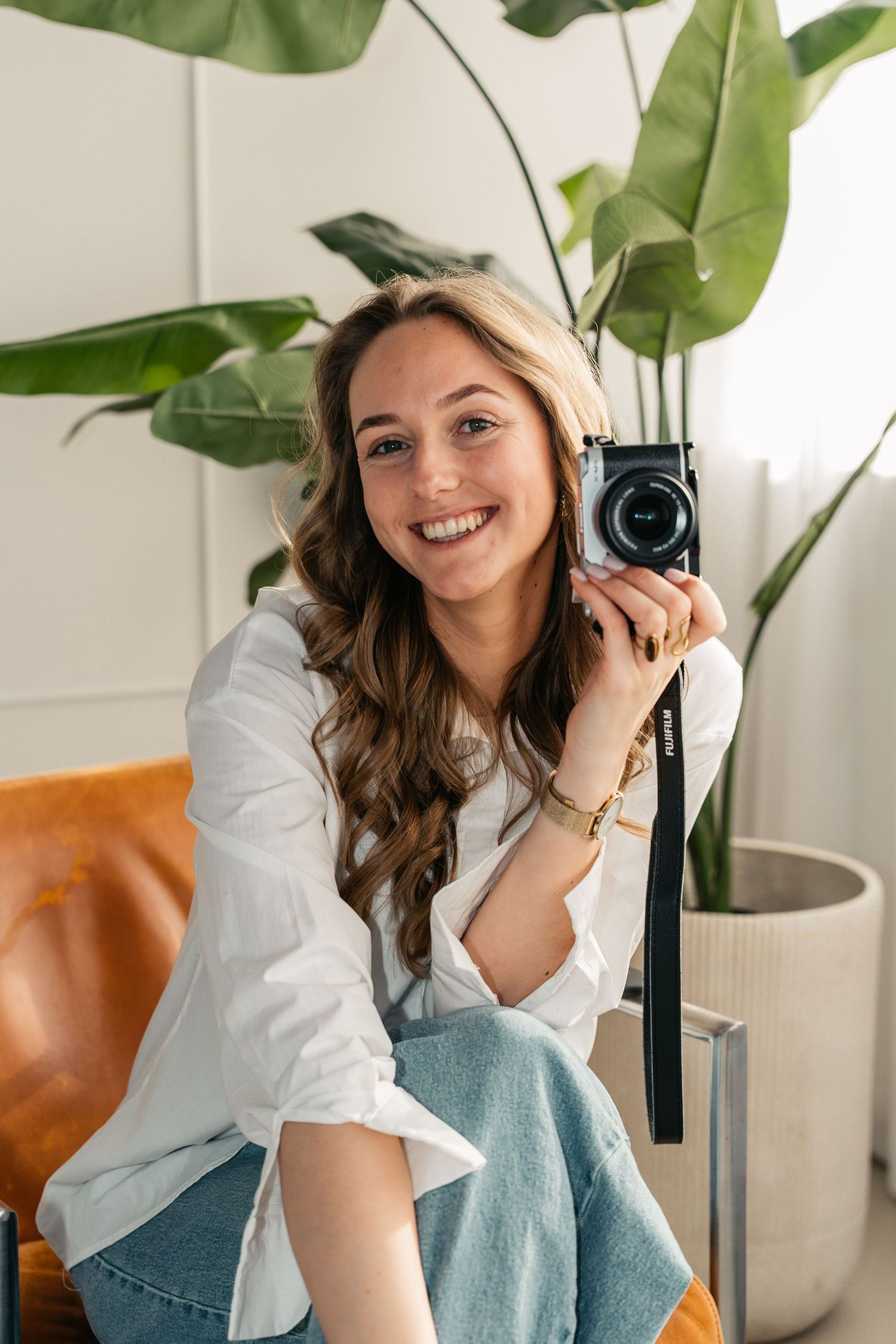 Smiling young woman holding a vintage camera, wearing white shirt and jeans, sitting near a large tropical plant indoors.
