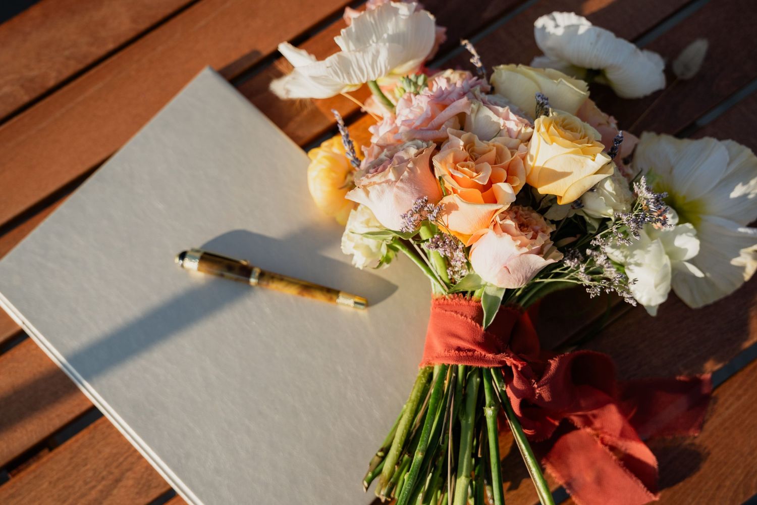 A bridal bouquet of peach and white roses tied with red ribbon beside a guest book and pen on a wooden table.