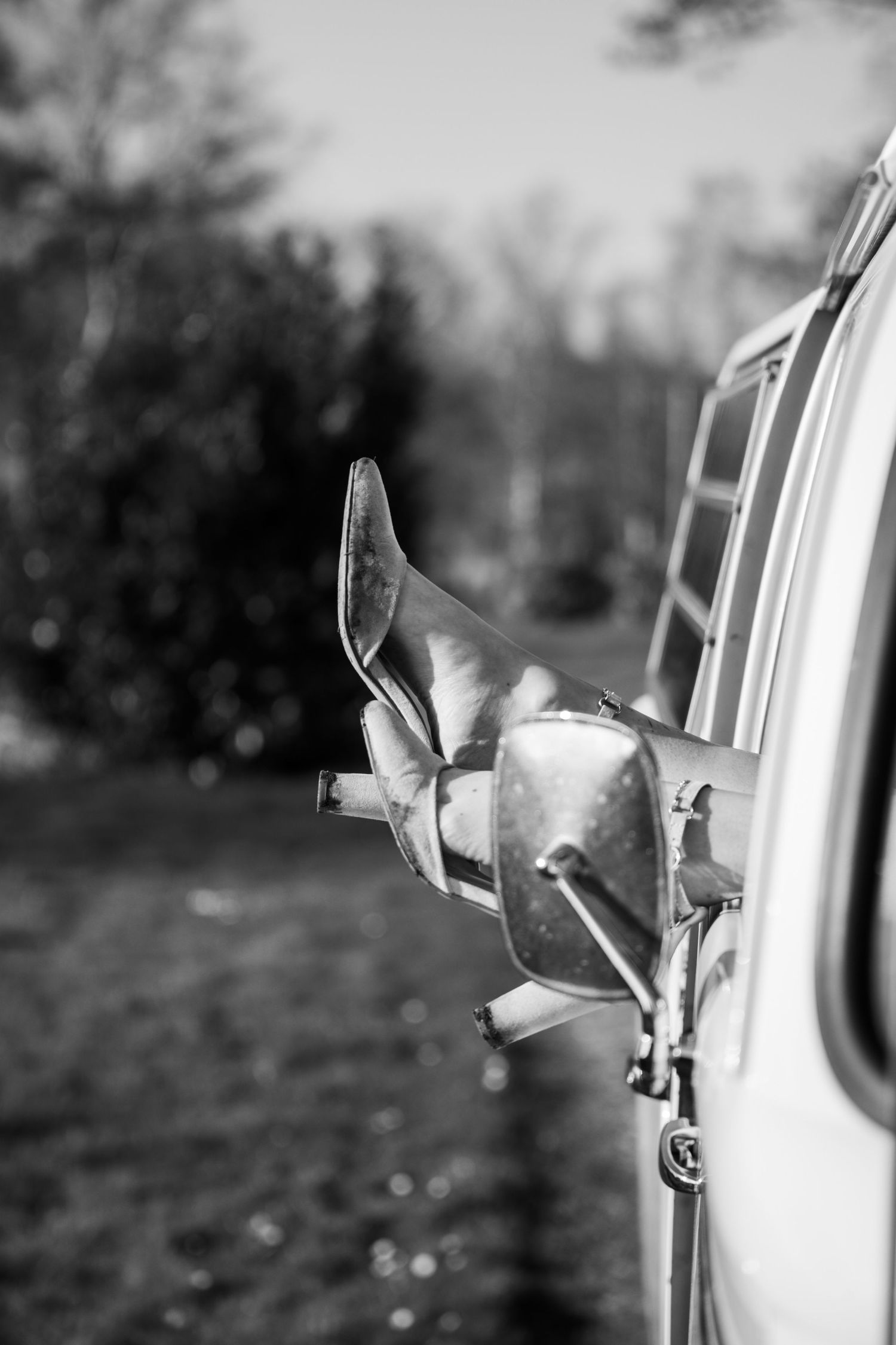 Black and white photo of high heel shoes hanging out of a vintage car window in a rural setting.