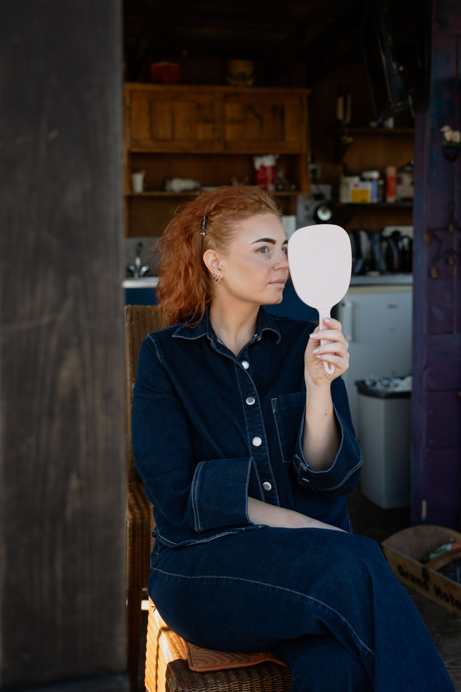 Red-haired woman in denim jumpsuit sitting on wicker chair holding white hand mirror in rustic kitchen setting.