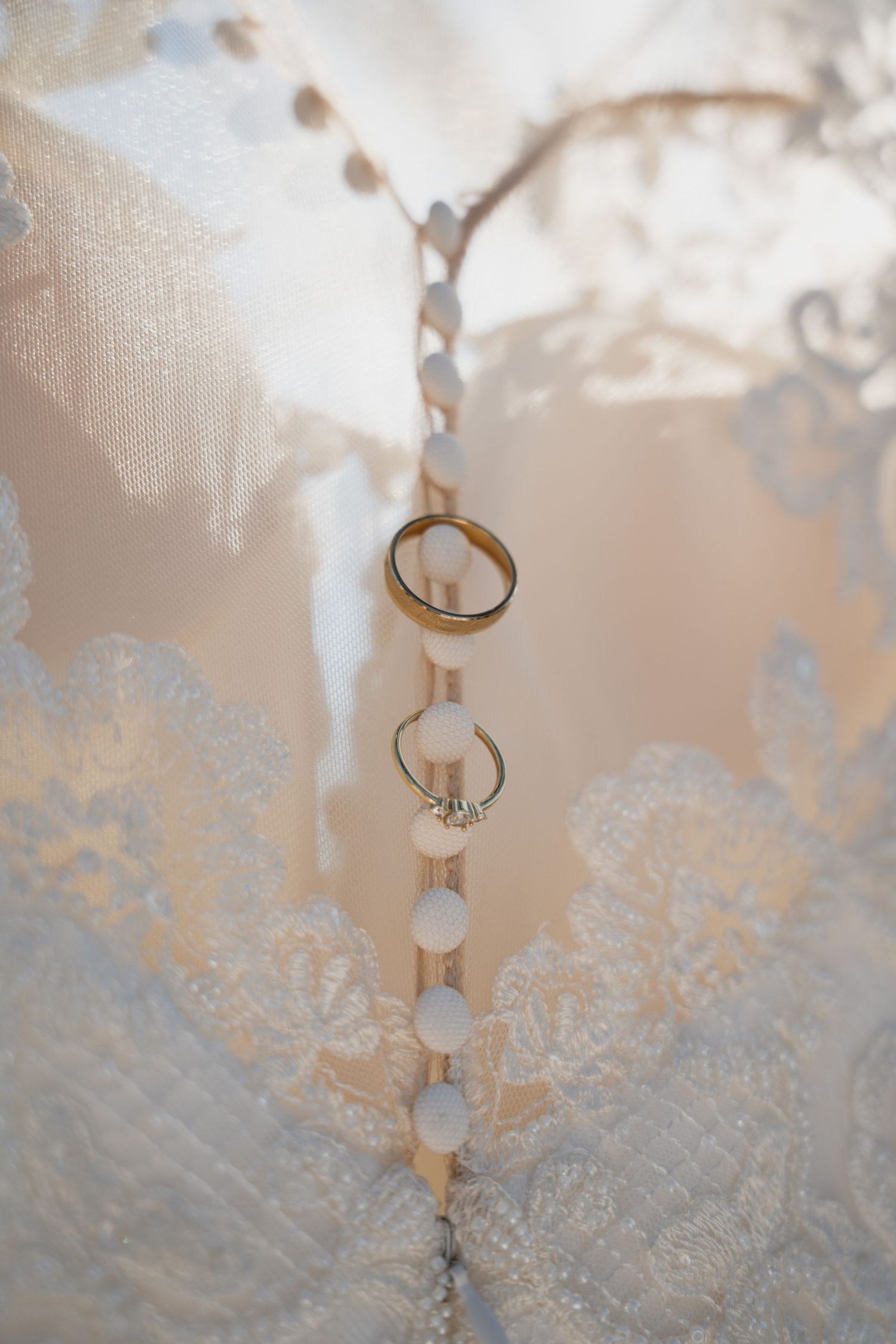 Two gold wedding bands and a diamond engagement ring hanging on pearl buttons of a white lace bridal gown.
