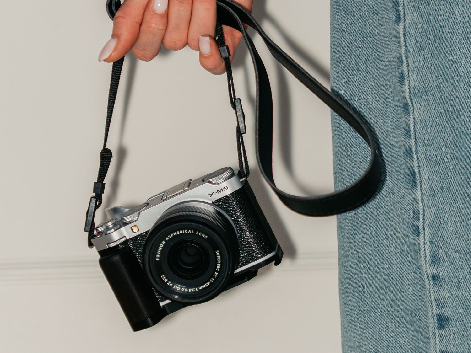 A hand holds a silver and black Fujifilm mirrorless camera with a black leather strap against a neutral background.