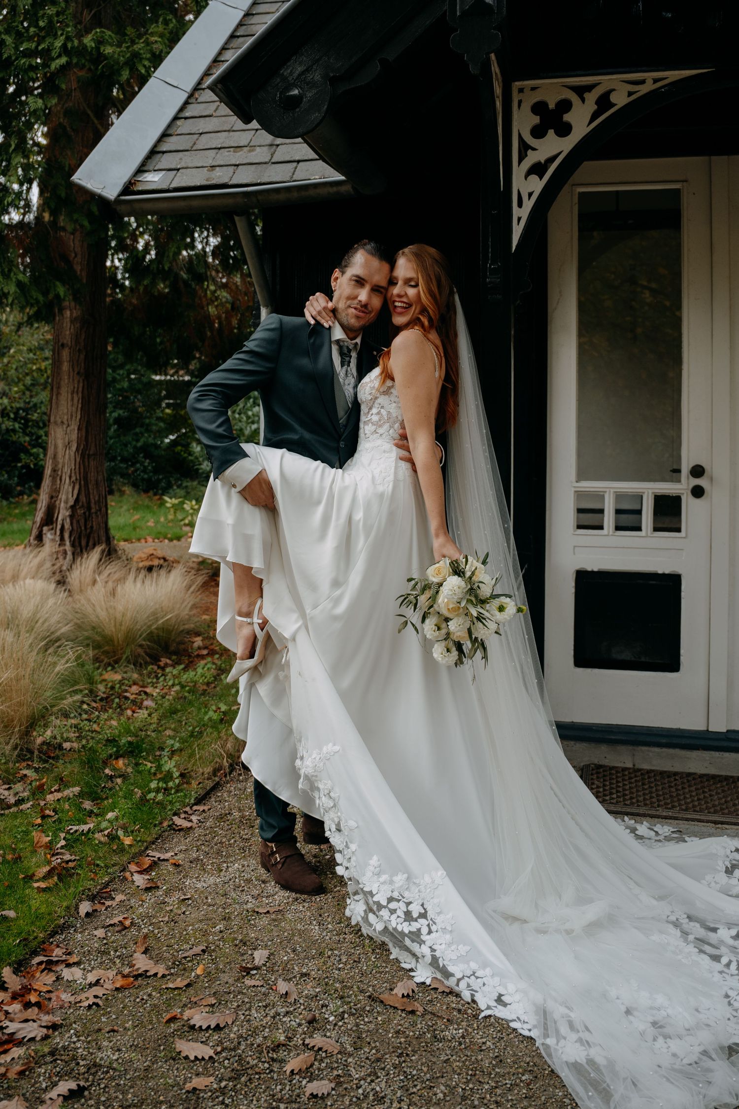 Couple in wedding attire posing together on gravel path near white cottage.