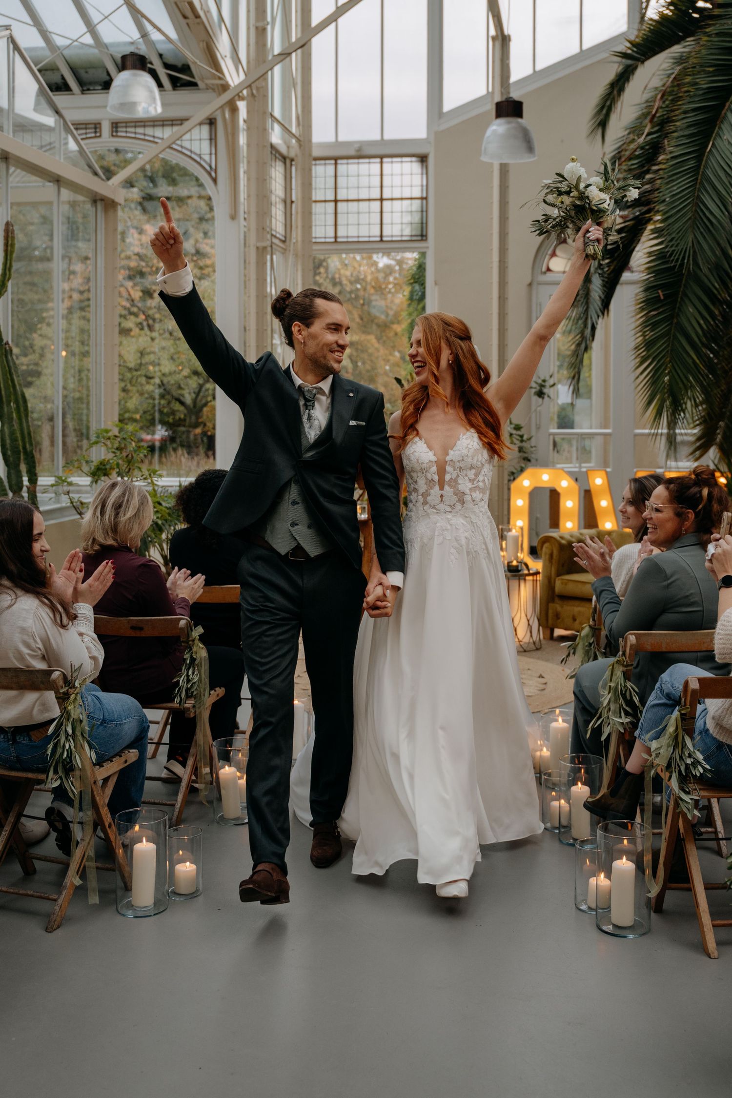 Wedding guests celebrating and cheering in a bright conservatory venue with candles and palm plants.