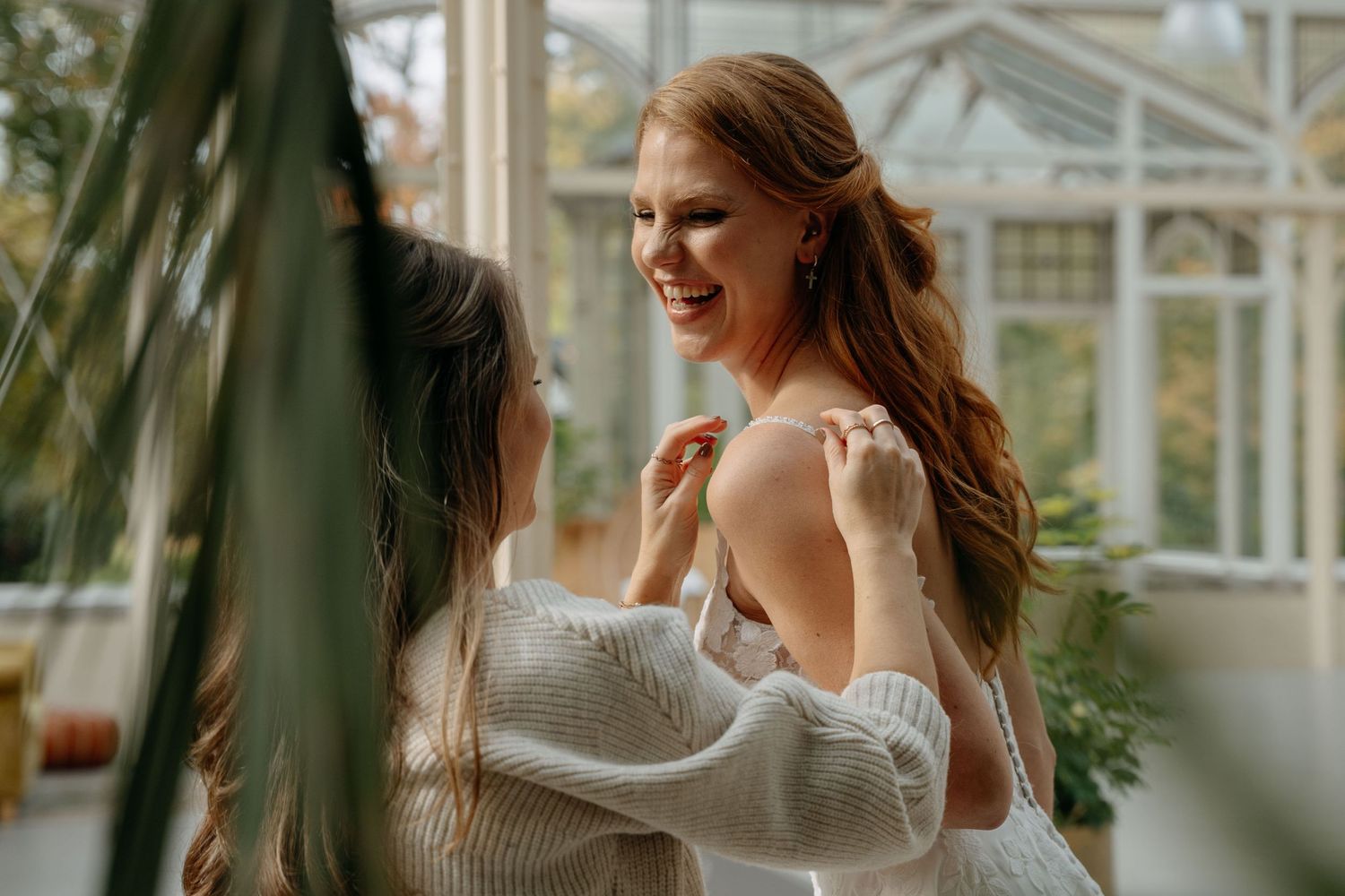 Person in white wedding dress smiles joyfully near greenhouse windows in natural lighting.