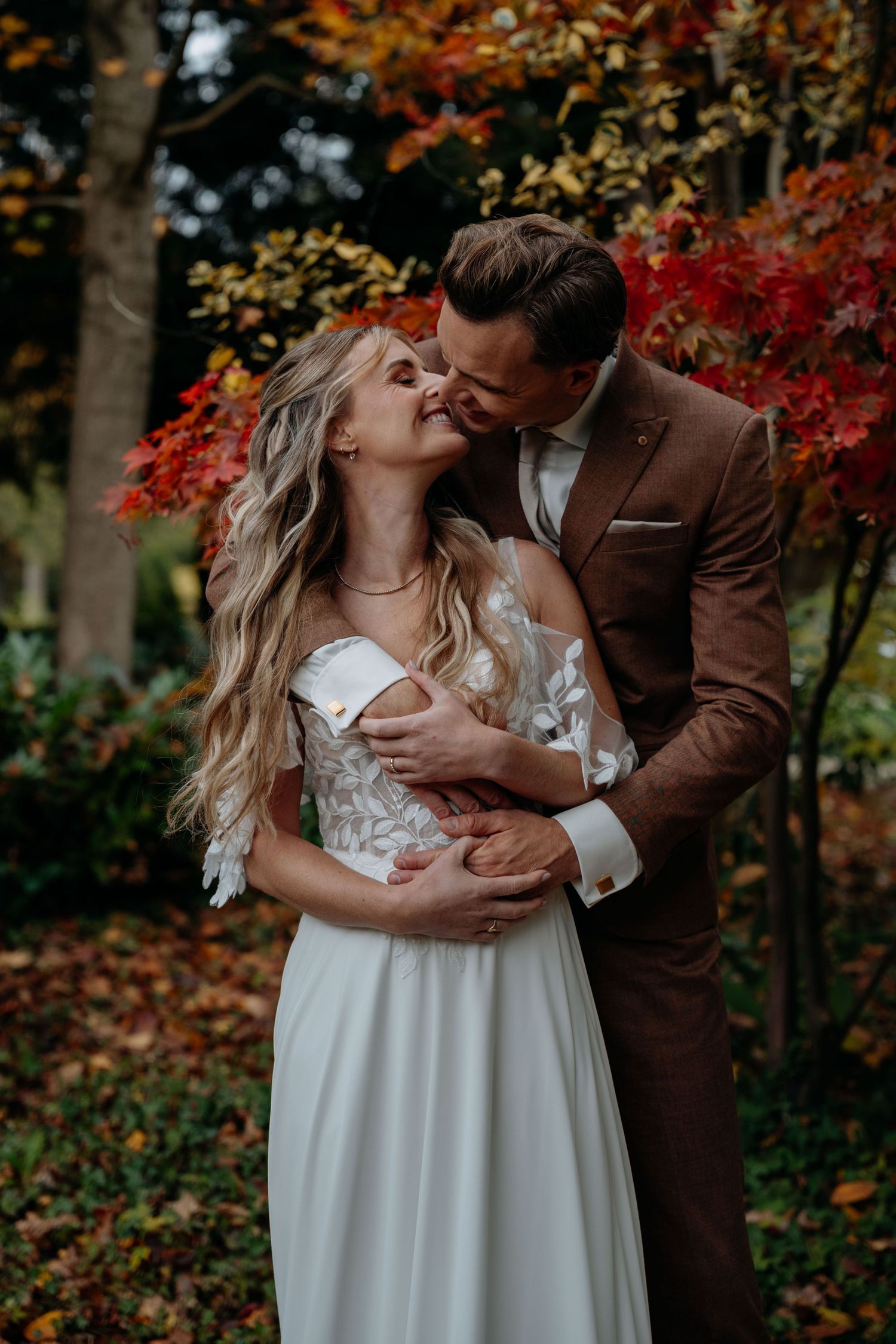 A romantic wedding couple embraces in front of vibrant autumn foliage wearing a white gown and brown suit.