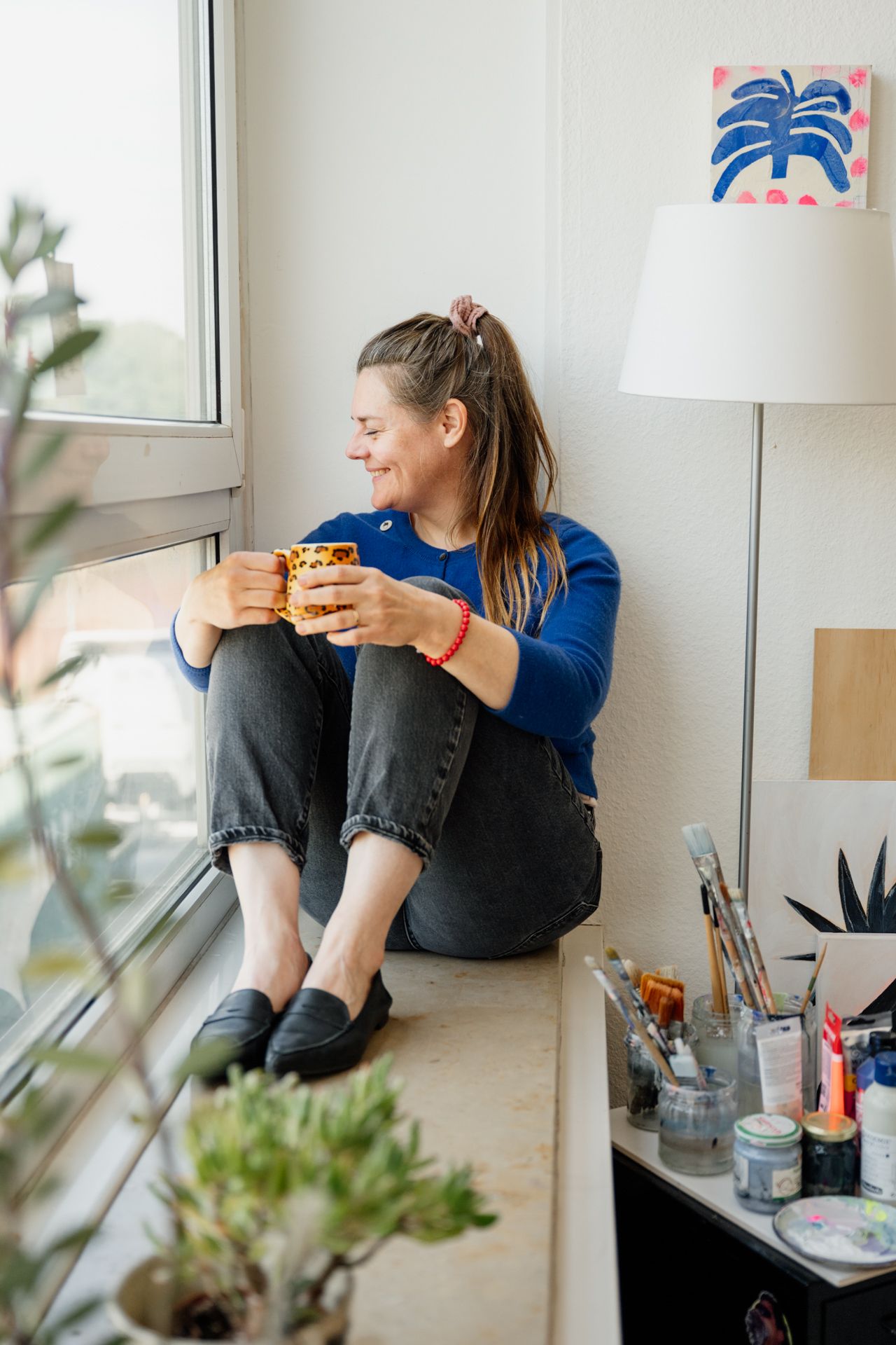 Künstlerin in ihrem Atelier sitzend und kaffeetrinkend auf Fensterbank