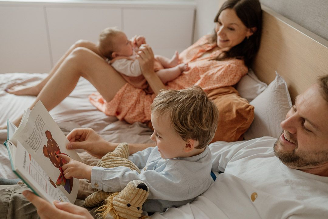 eine Familie mit Baby und Kleinkind sitzt auf dem Bett und liest gemeinsam während Homestory