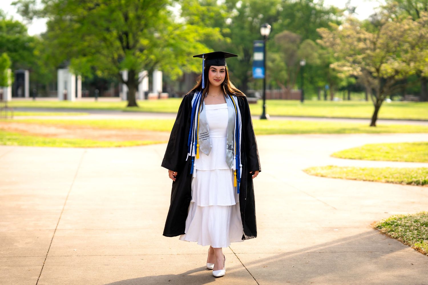 Graduate in black cap and gown over white dress walks on campus path during outdoor graduation photo session.