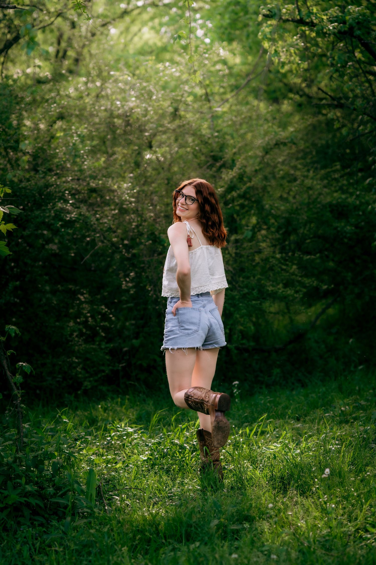 Young woman in white top, denim shorts, and brown boots smiling in a lush green forest setting.