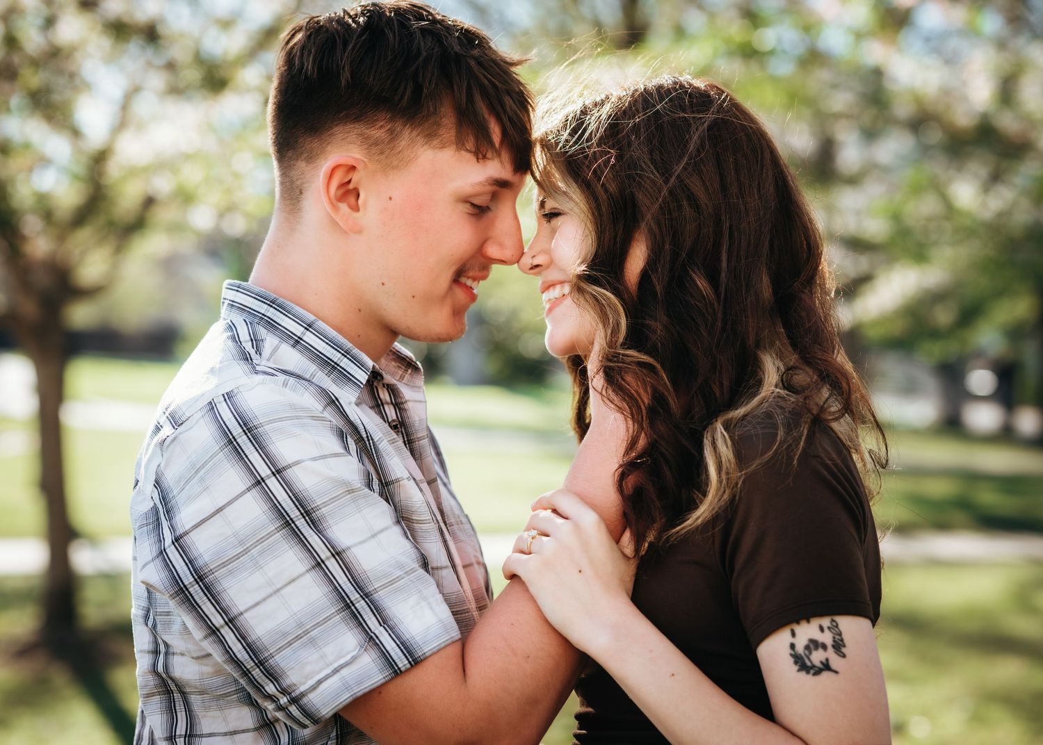 Romantic couple embracing outdoors in a sunny park, touching foreheads affectionately, woman with tattoo visible.
