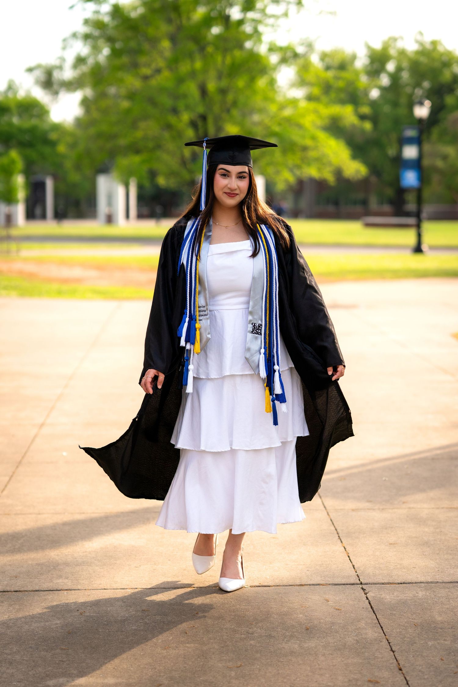 Graduate in black cap and gown over white dress posing on campus walkway with trees in background.