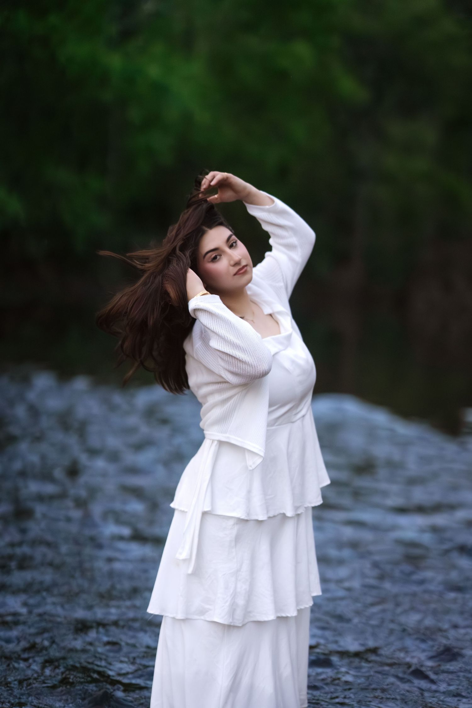 Young woman in white tiered dress standing in river, lifting flowing brunette hair with green forest background.