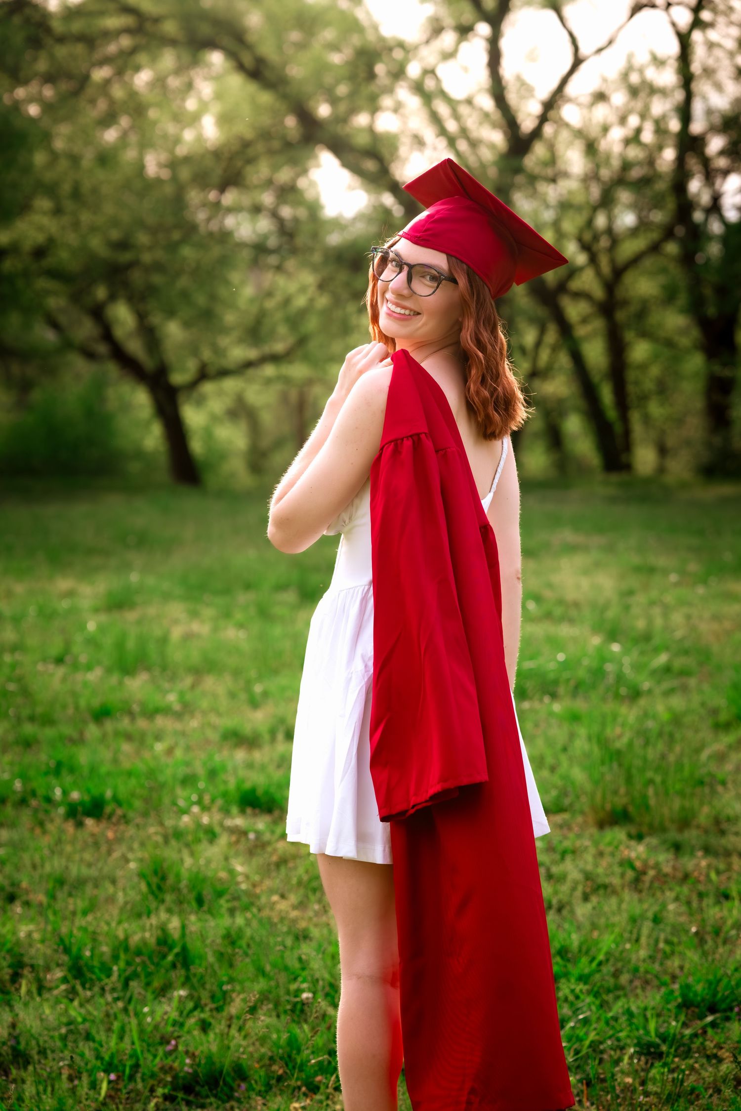 Smiling young woman in red graduation cap and gown holding her robe outdoors in a lush green park setting.