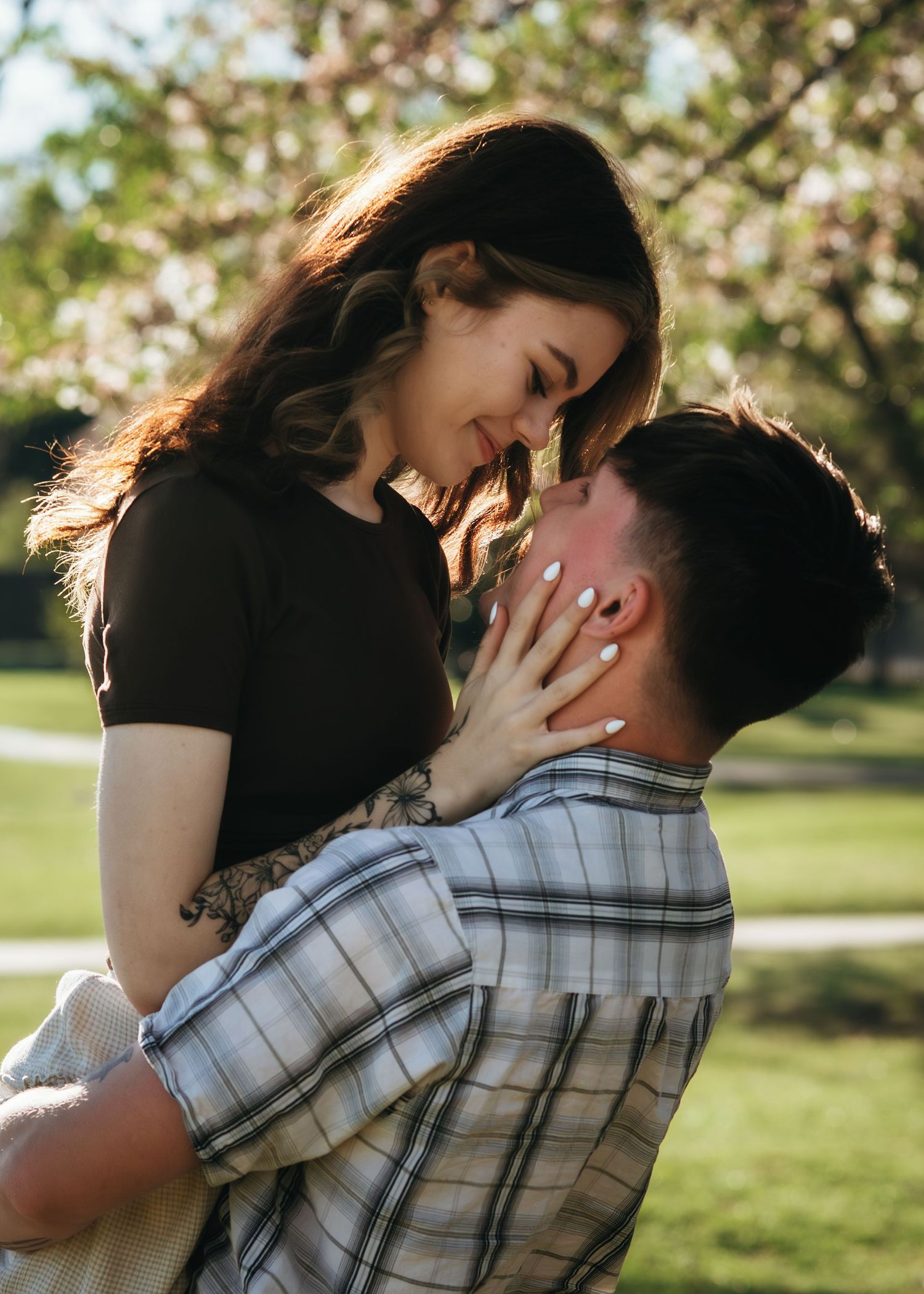 Romantic couple embracing outdoors, man lifting woman as they smile at each other near blooming trees.