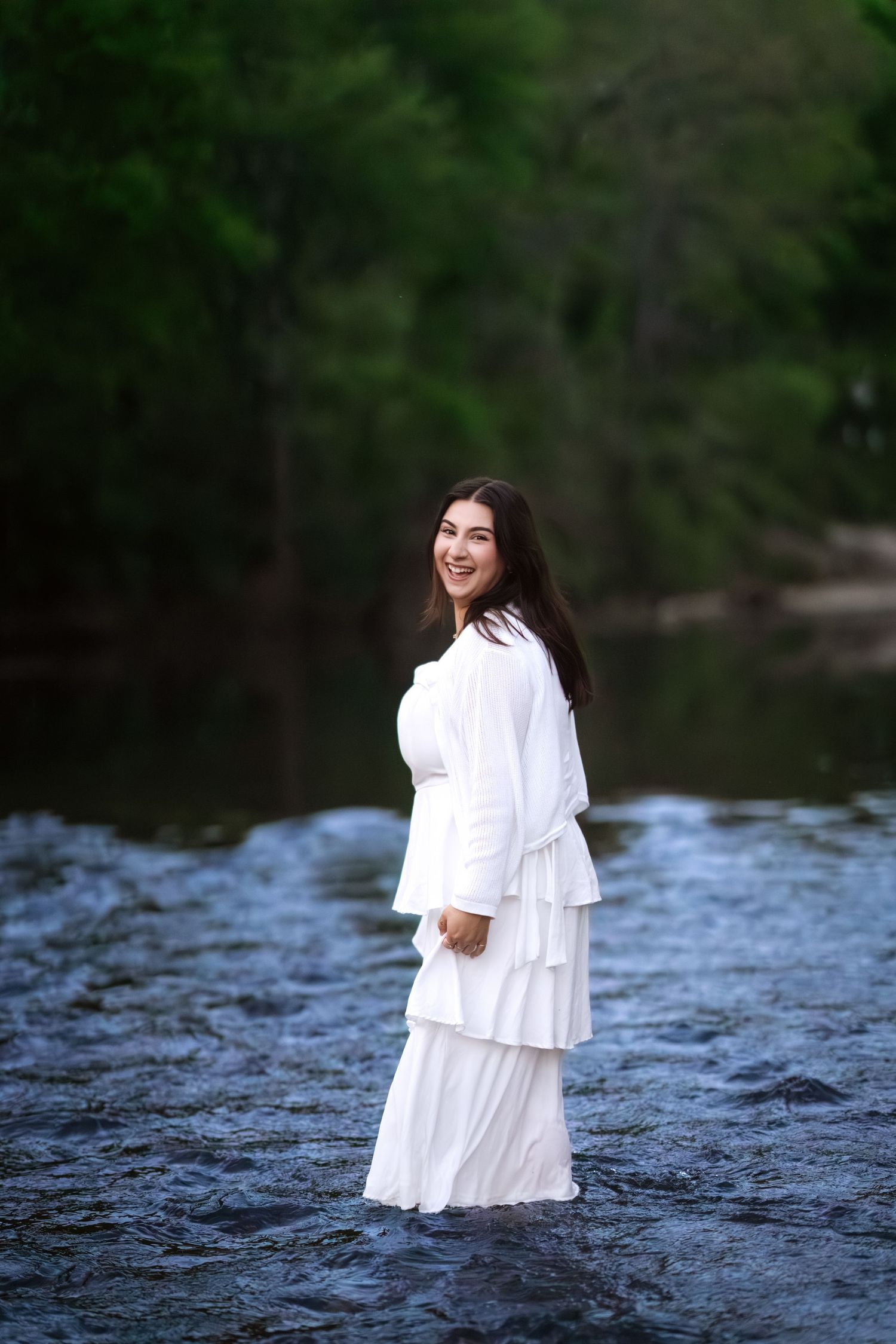 Smiling young woman in white tiered maxi dress standing in shallow river with lush green trees behind her.
