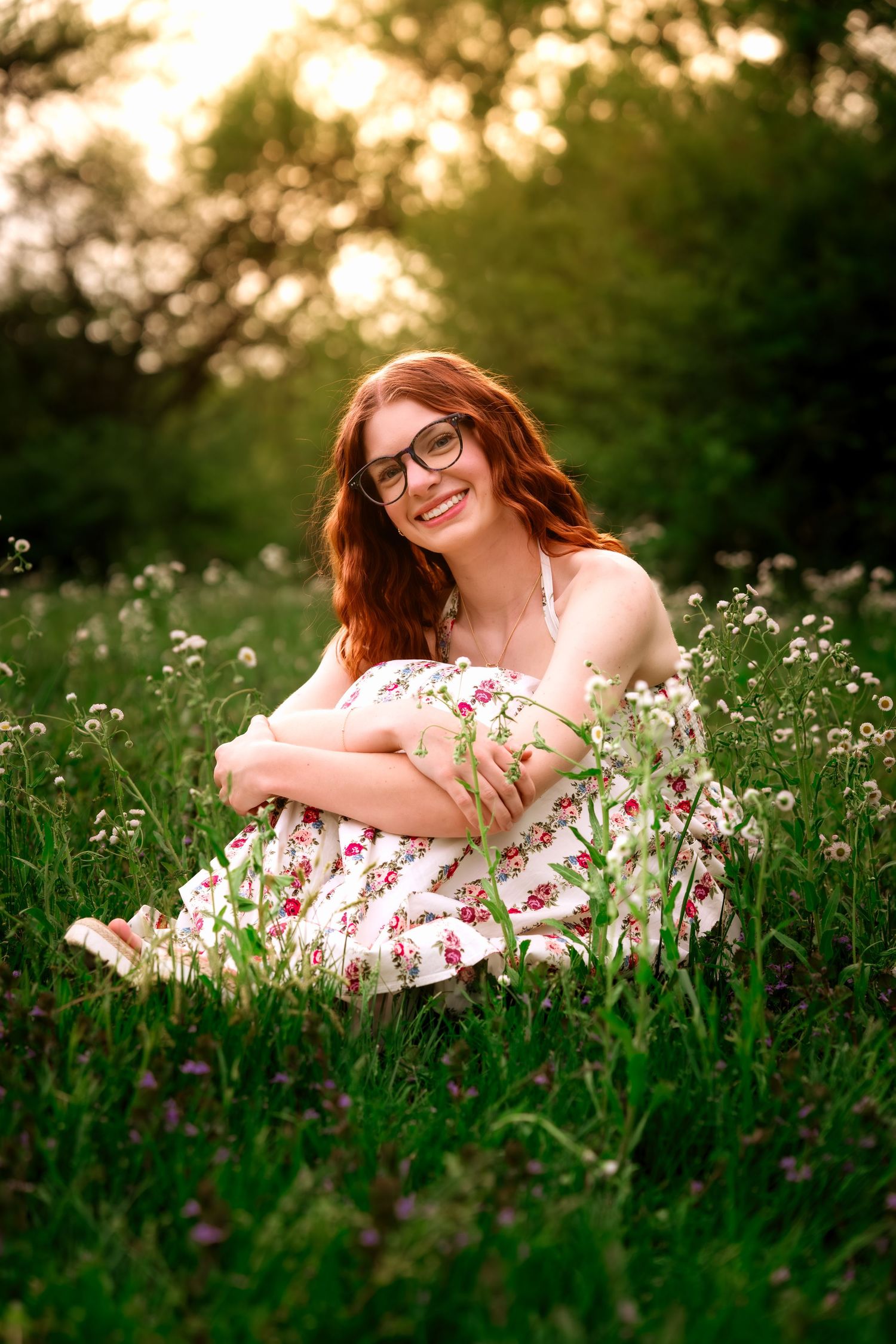 Smiling redhead woman in floral dress sitting in a wildflower meadow during golden hour sunset.