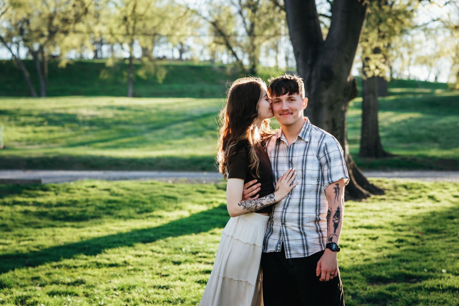 Couple embracing in a sunny green park, woman kissing man on cheek, both with tattoos, trees in background.