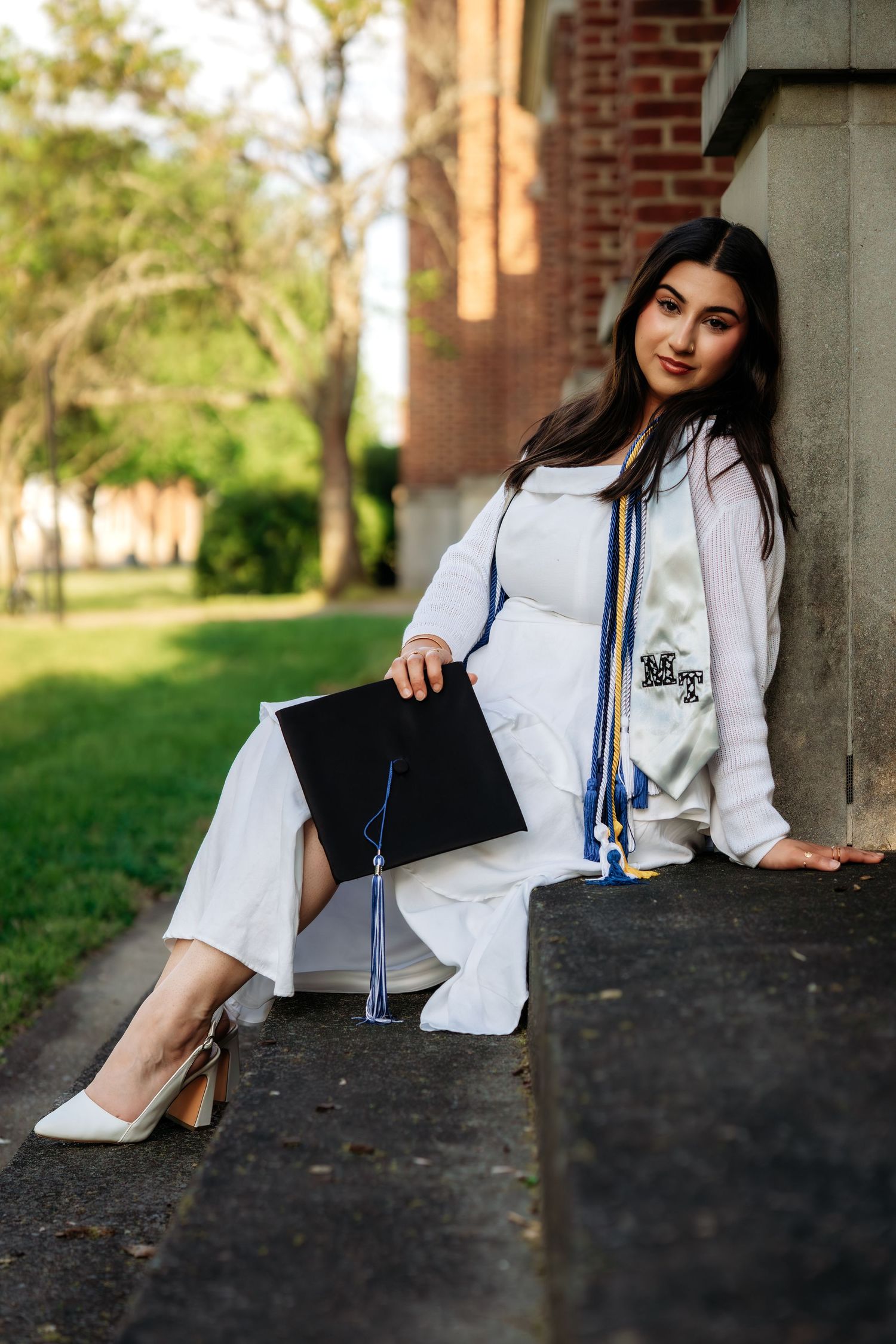Female graduate in white dress and honor cords sits on stone steps holding black mortarboard cap.