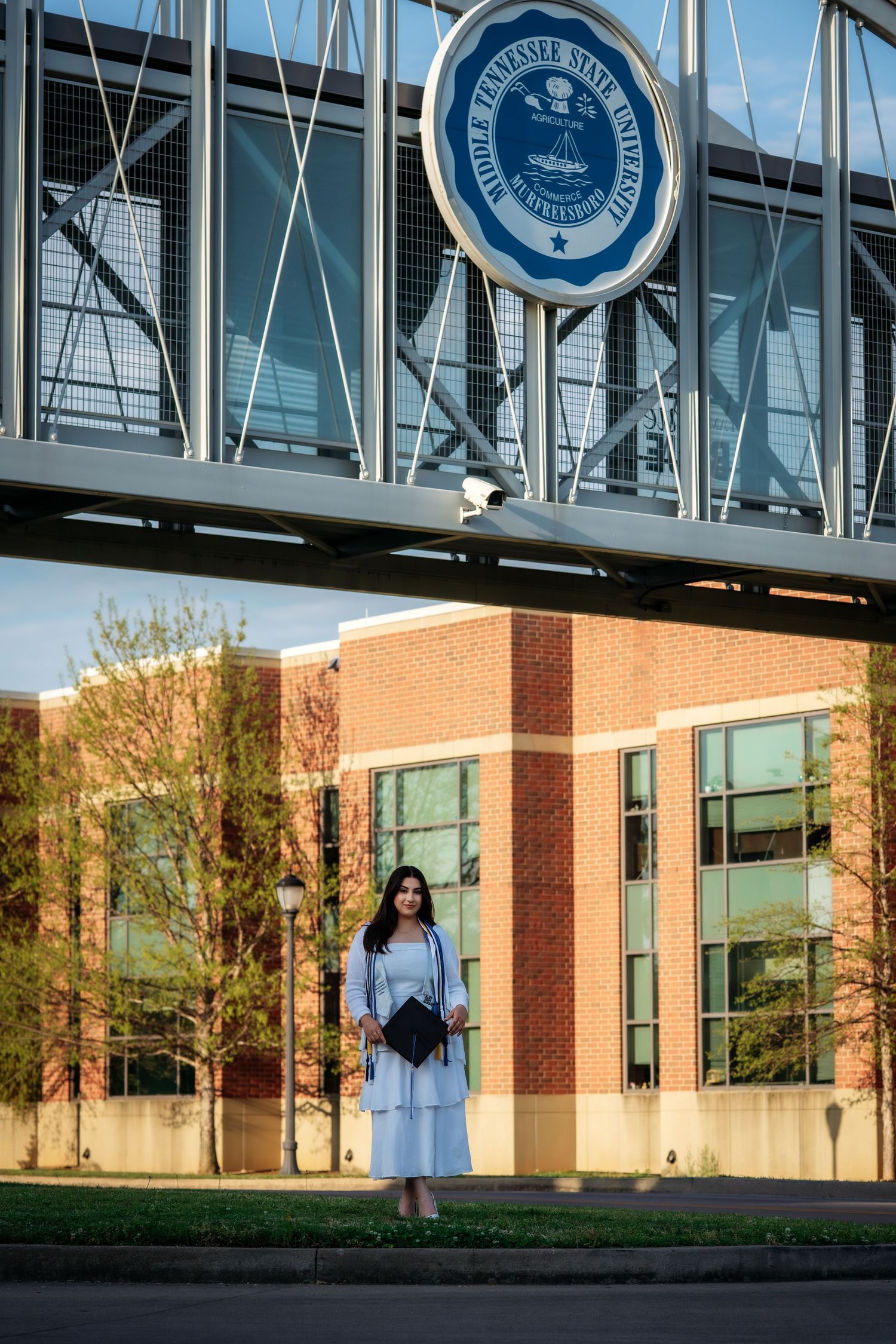 Graduate in white dress holding cap stands under Middle Tennessee State University seal bridge on campus.