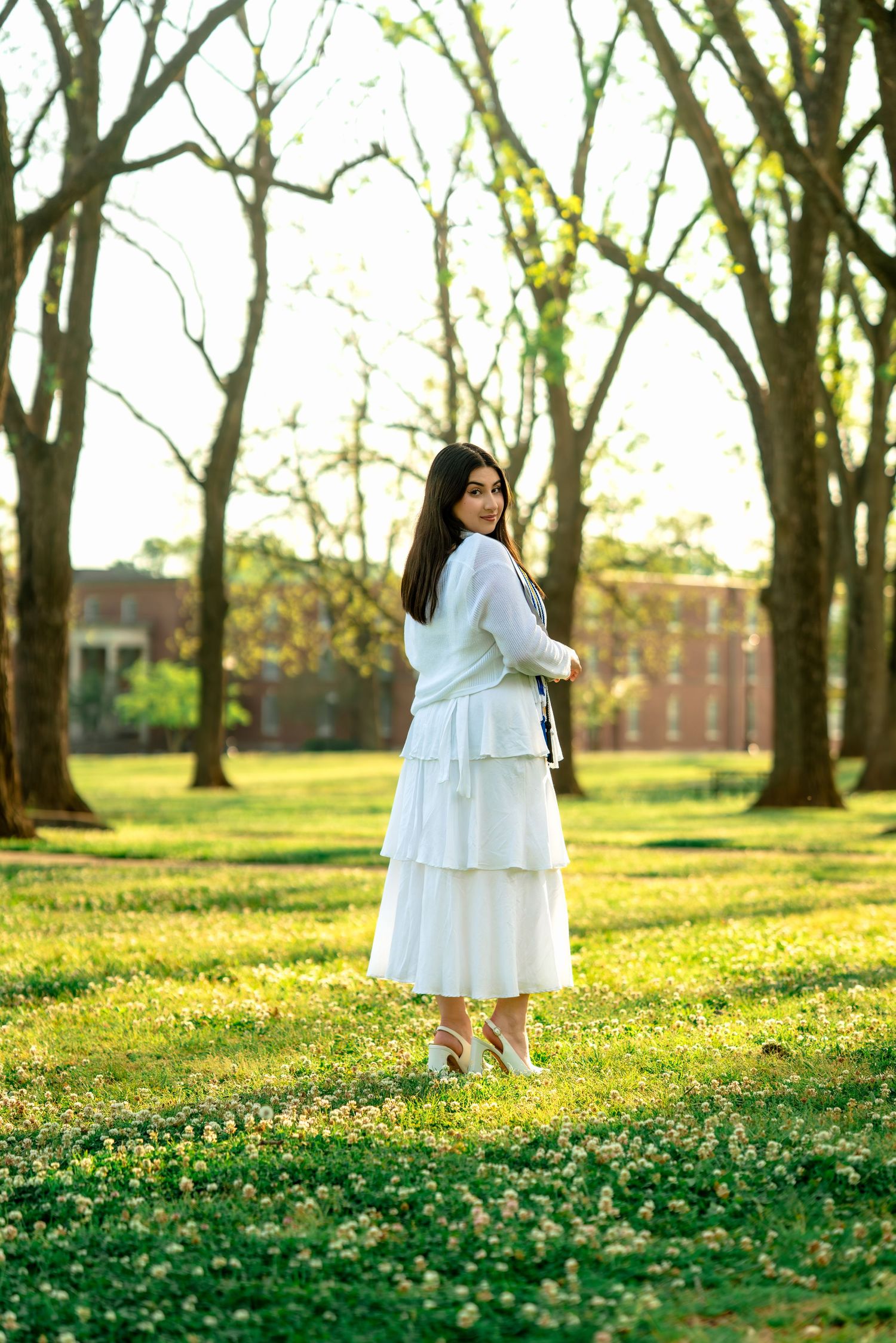 Young woman in elegant white tiered dress walks through sunlit tree-lined campus pathway in spring.