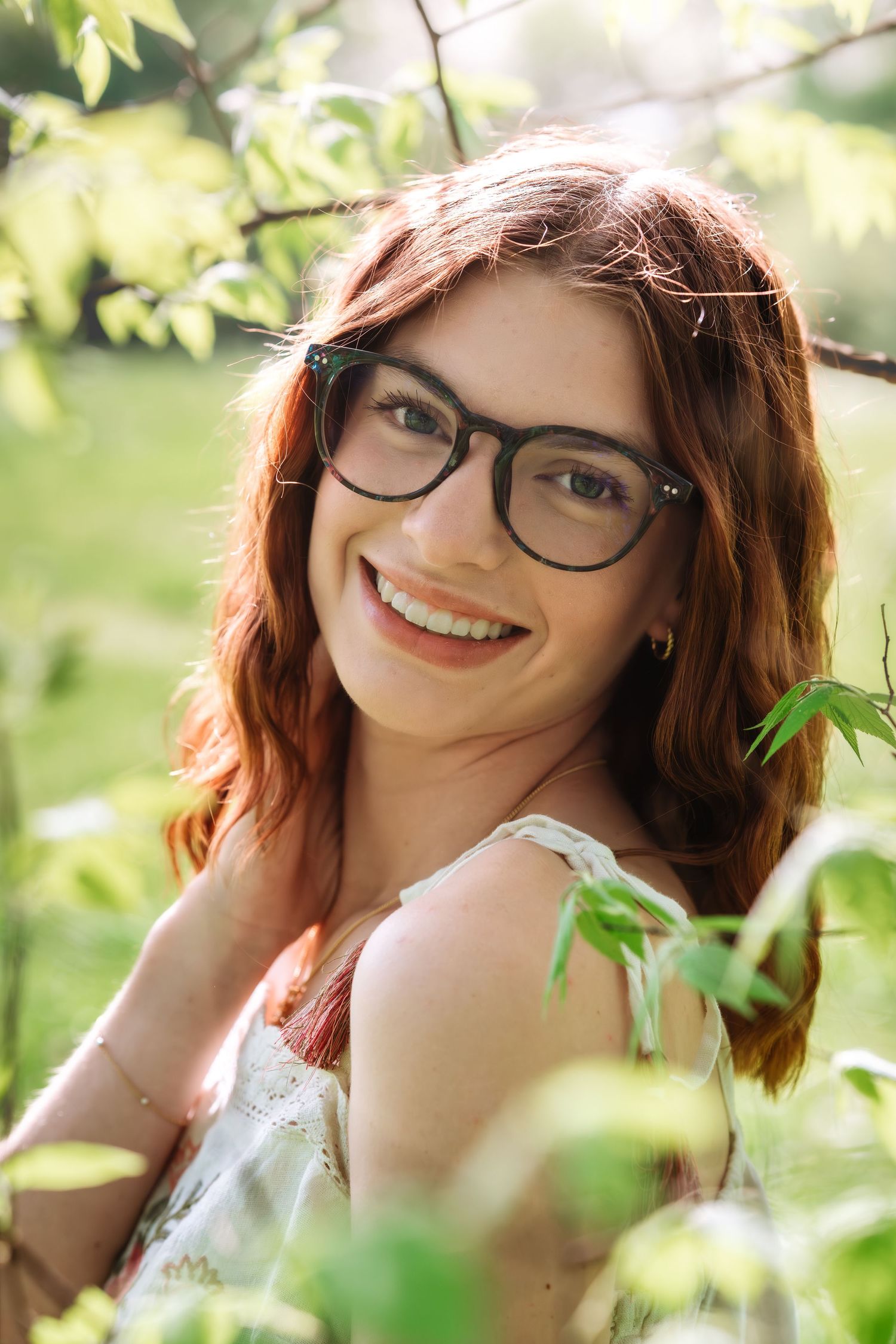 Smiling redhead woman wearing glasses surrounded by green leaves in a sunny outdoor setting.