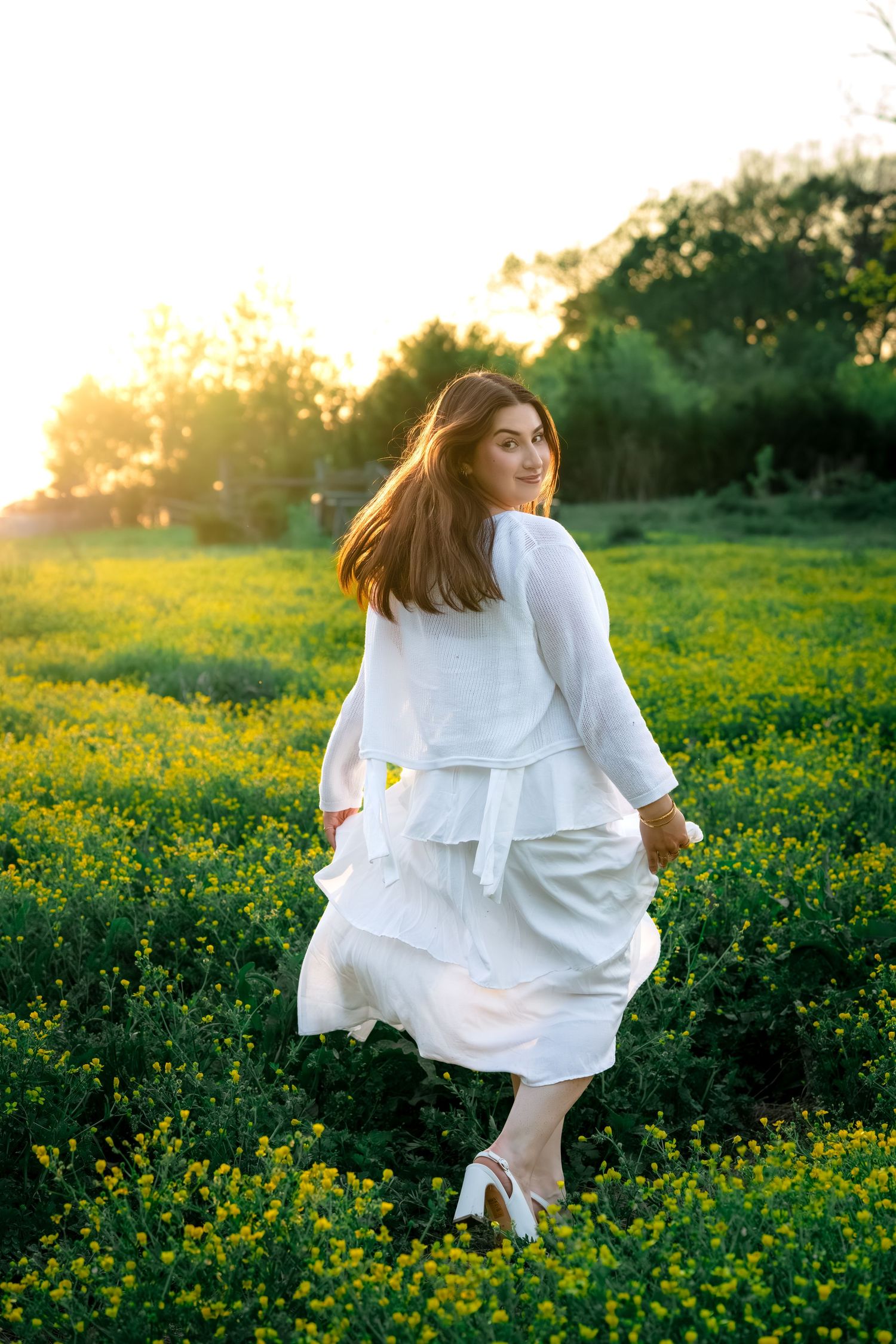 Woman in flowing white dress walking through yellow flower meadow at sunset.