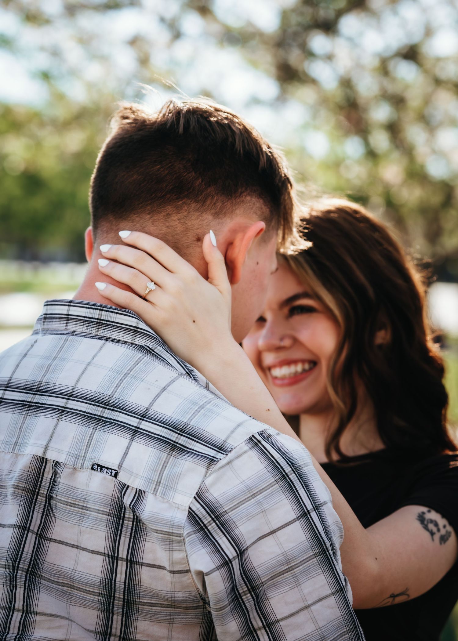 Smiling woman with tattoo and ring embraces man in plaid shirt outdoors in sunny park setting.