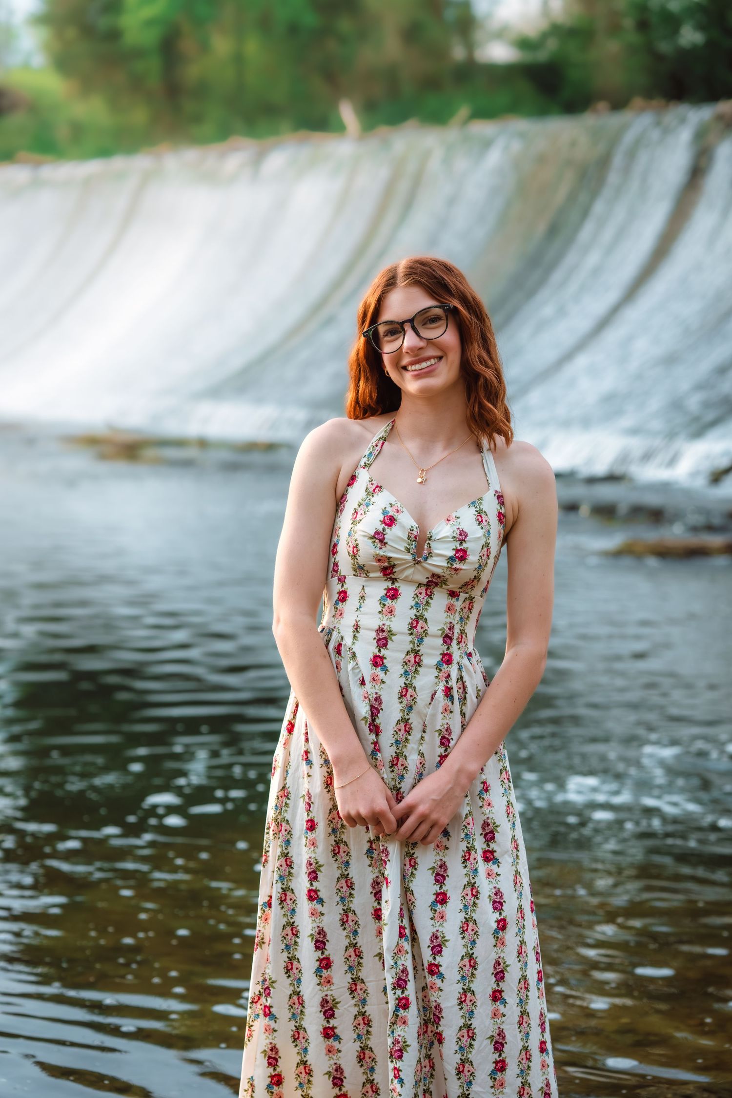 Smiling redhead woman in floral maxi dress standing in shallow water near a waterfall wearing glasses.