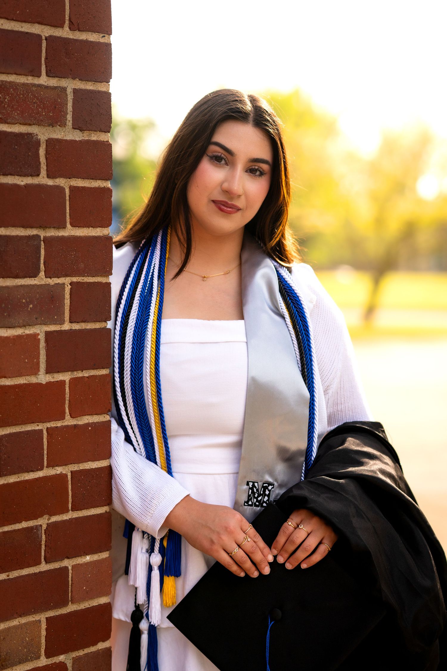 Young woman in white dress and blue graduation stole leans against brick wall holding cap and gown.